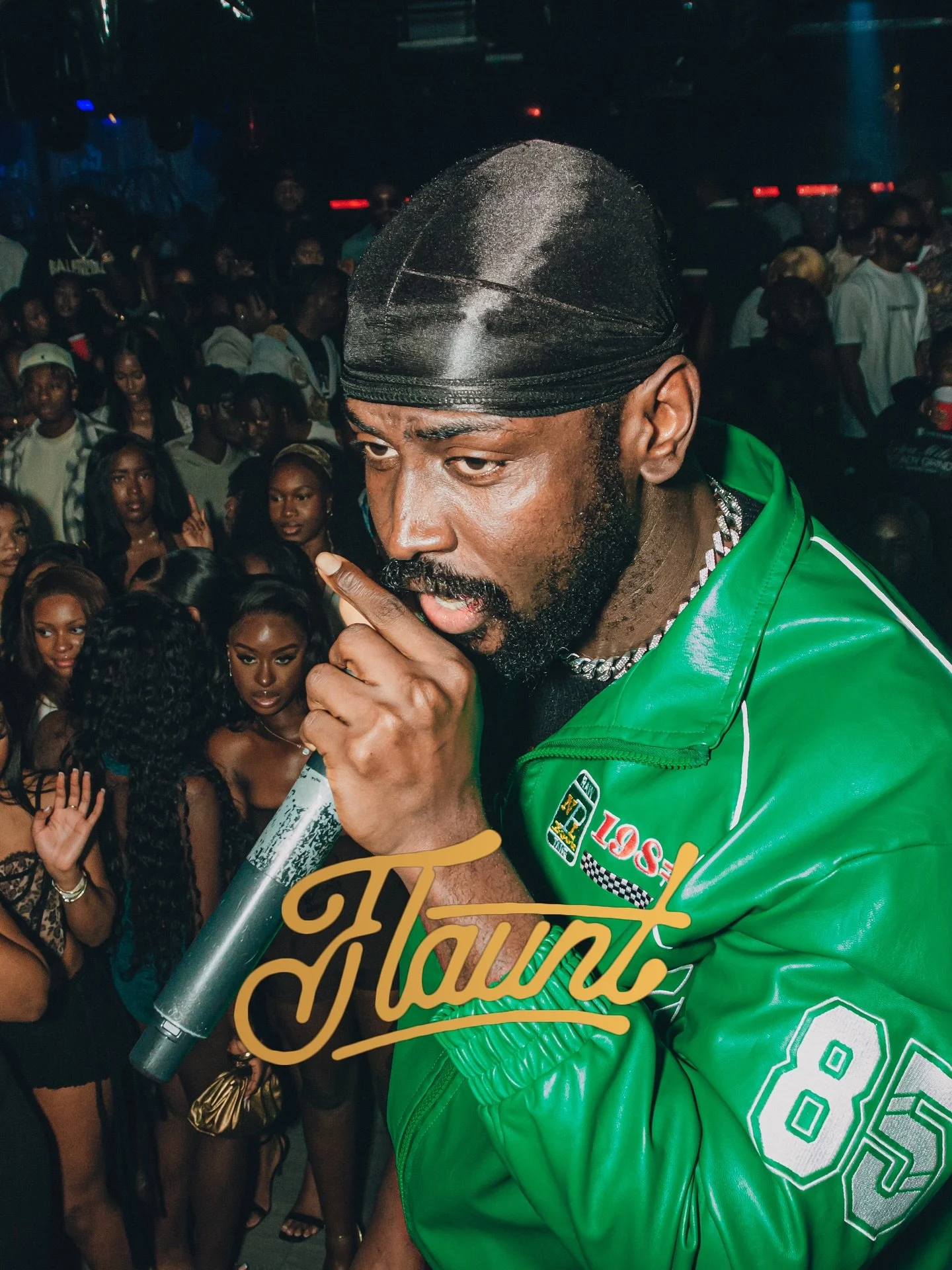 A man in a green jacket and black durag holding a microphone on stage, with a crowd of young women and men behind him at a nightclub or concert.