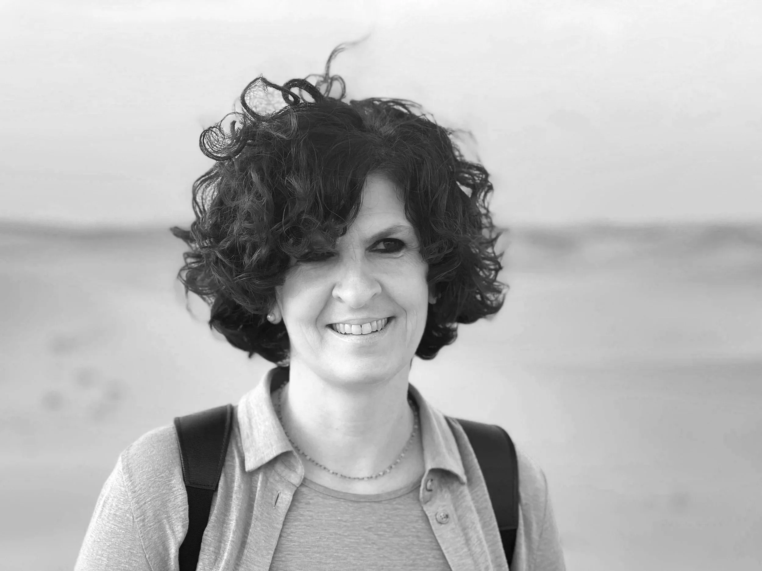 A woman with curly dark hair smiling outdoors at the beach, wearing a purple shirt and backpack.