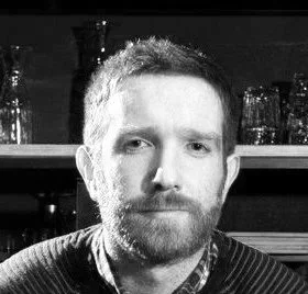 A man with red hair and a beard, posing indoors with shelves of glasses in the background.