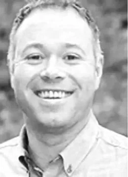 A man smiling outdoors with a blue collared shirt and green blurred background.
