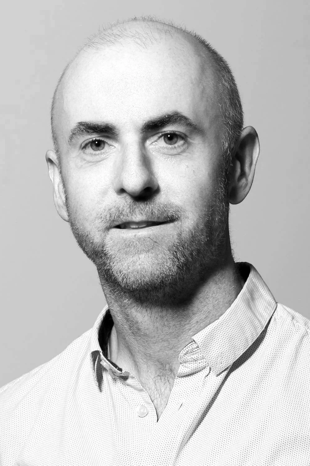 Close-up portrait of a middle-aged man with a shaved head, gray beard, and light-colored eyes, wearing a white collared shirt, against a plain gray background.