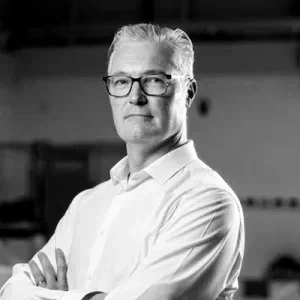 A man with gray hair and glasses wearing a white shirt, standing with arms crossed in an indoor industrial or warehouse setting.