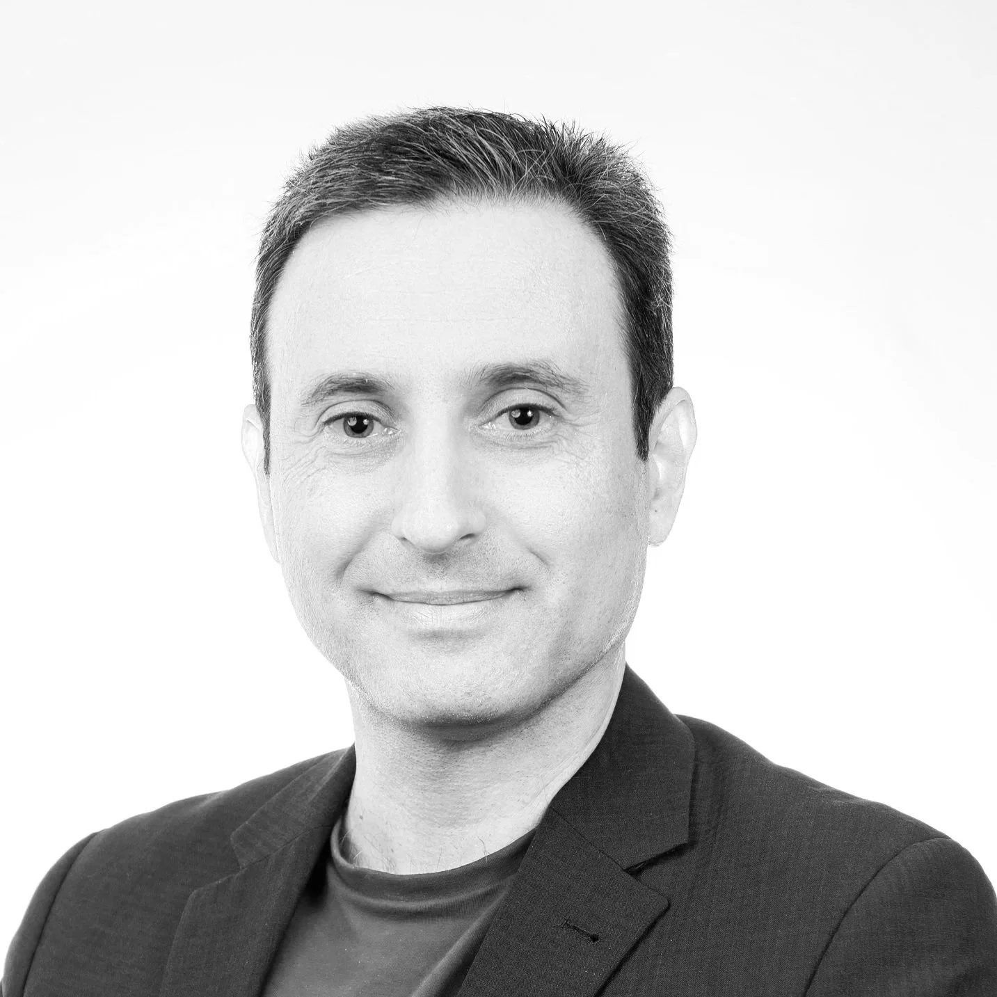 Portrait of a man with short dark hair, blue eyes, wearing a dark blazer and maroon shirt, smiling against a plain white background.