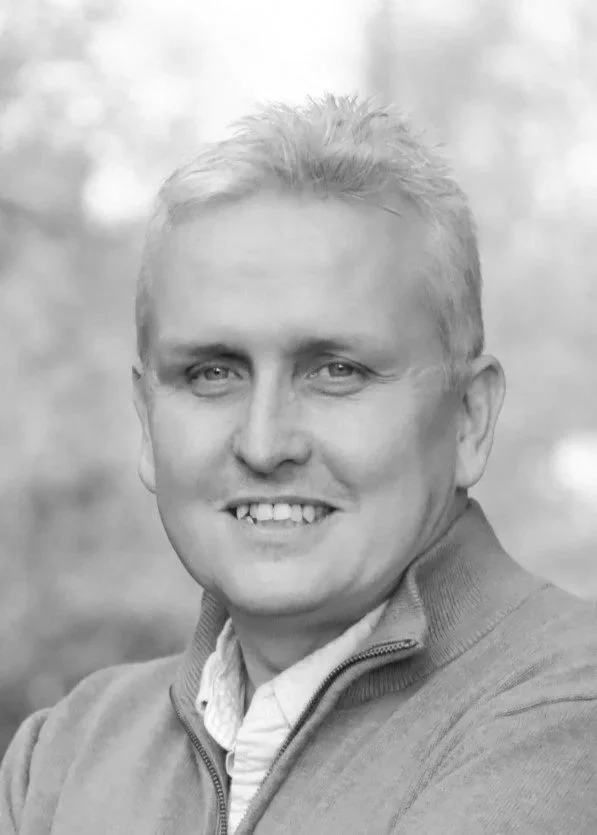 A man with short gray hair and blue eyes smiling outdoors, wearing a light-green zip-up jacket over a white collared shirt.