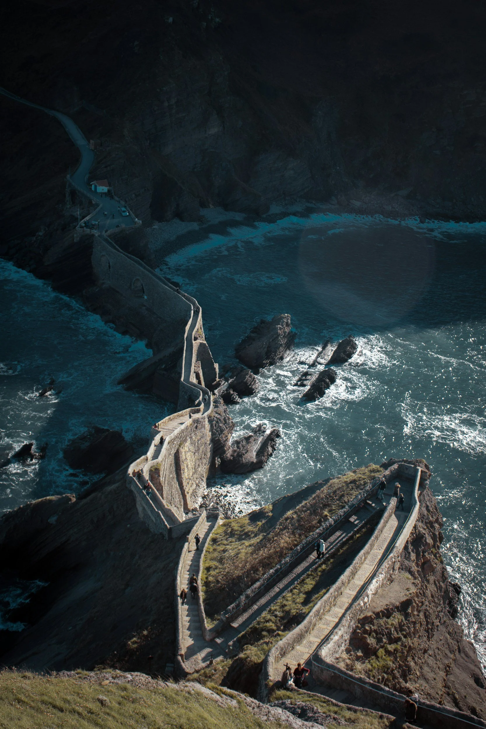 Vue aérienne d'une falaise avec un chemin sinueux bordant l'océan et menant à un petit bâtiment, avec des rochers dans l'eau en contrebas.
