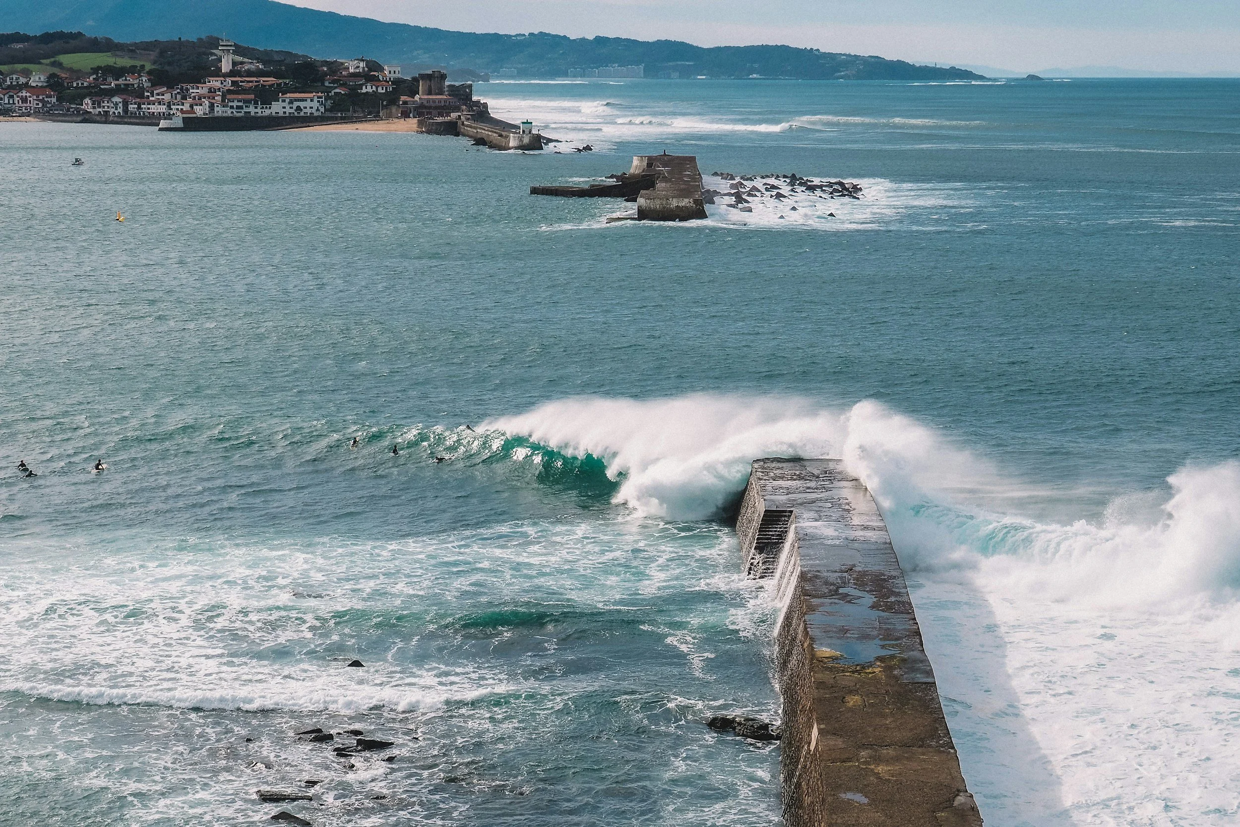 Vue de l'océan avec des vagues géantes frappant un brise-lames en béton, avec un groupe de surfeurs à proximité et une ville en arrière-plan sur la côte.