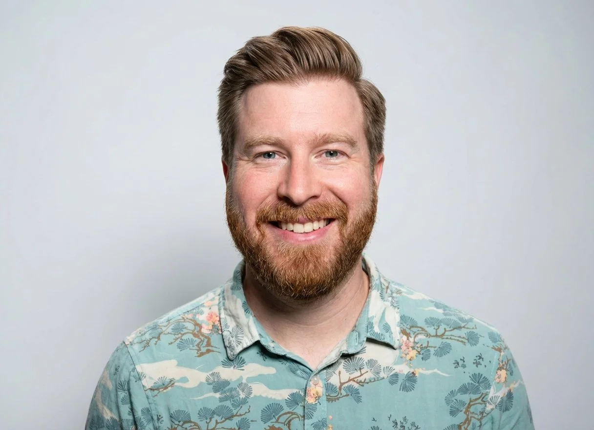 A smiling man with a beard and blonde hair, wearing a light blue patterned shirt, against a plain gray background.