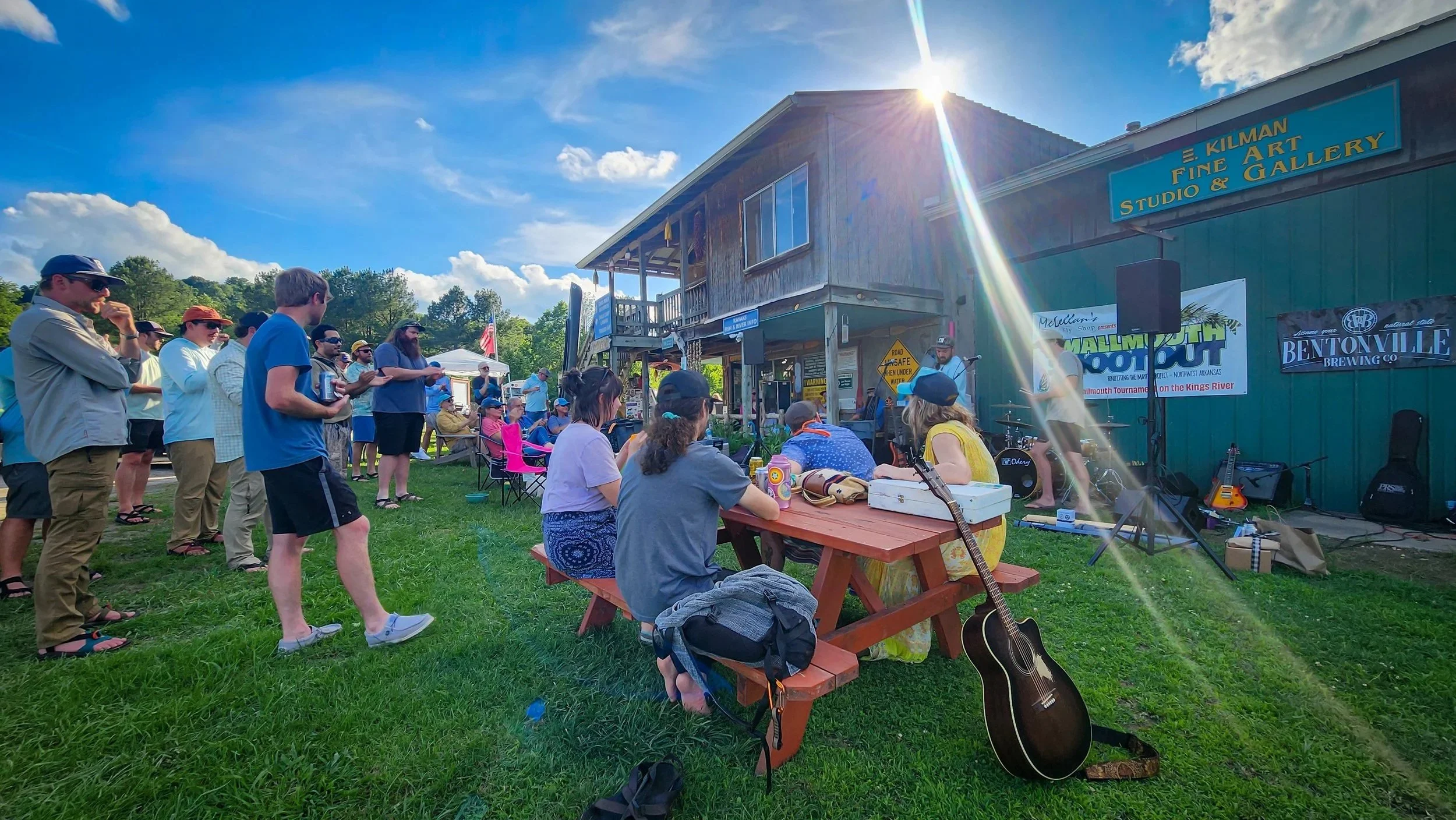 People gathered outdoors on a grassy area watching a live music performance on a stage connected to a green building with a blue sign. The sun is shining brightly in a clear sky at Kings River Outfitters.