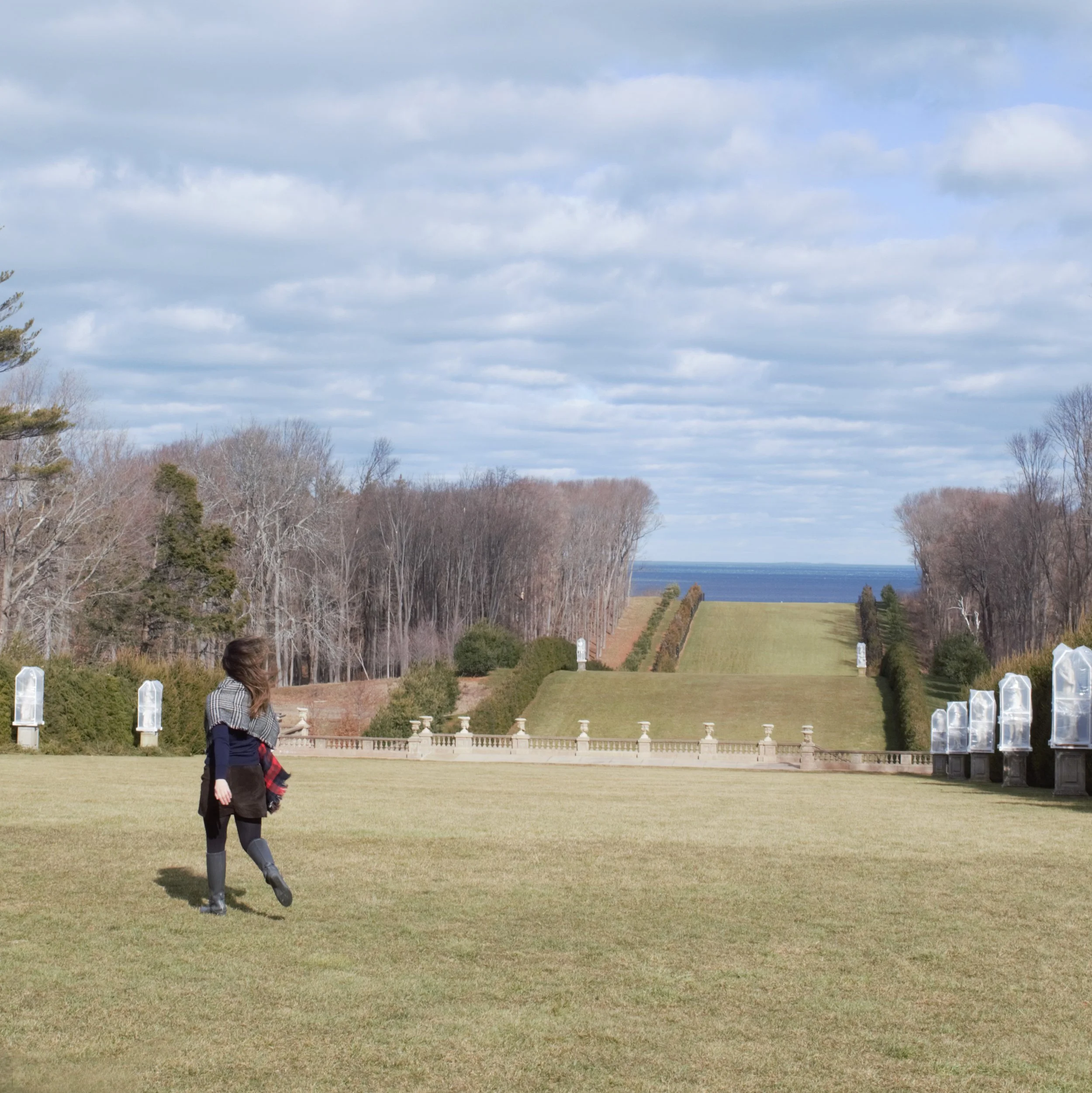 A person walking on a large open lawn facing a hill with a staircase, trees on both sides, and the ocean visible in the distance under a partly cloudy sky.