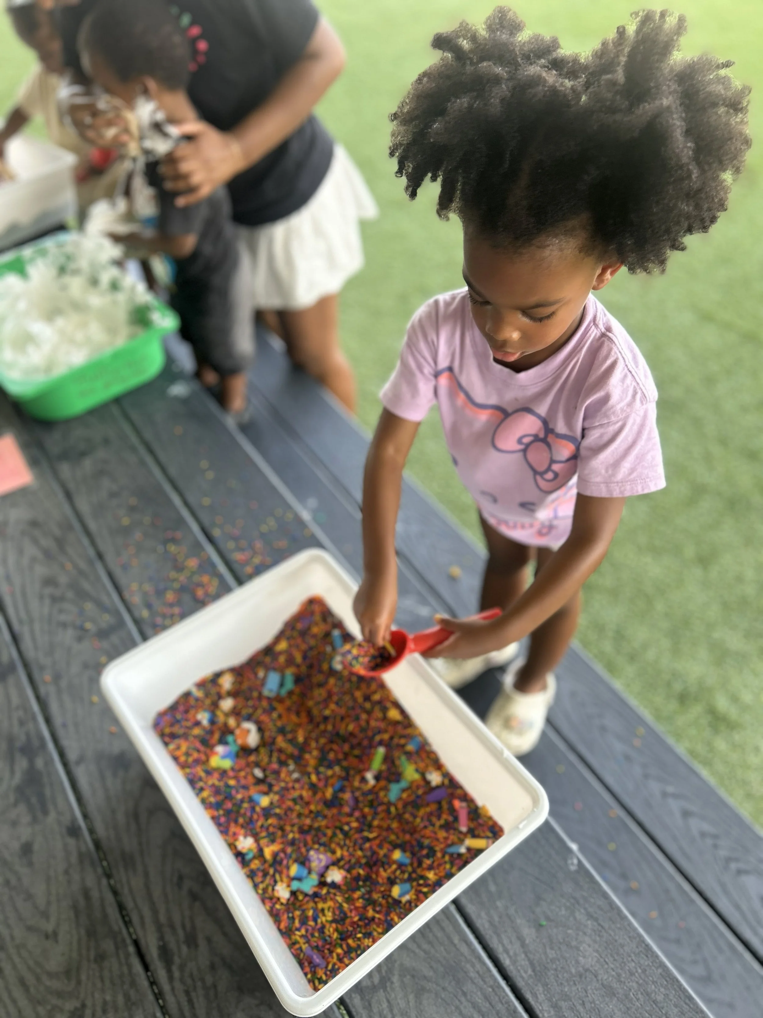 A young girl with curly hair wearing a pink Hello Kitty T-shirt is using a small scoop to pick colorful sprinkles from a white container. Other children and an adult are in the background on a wooden table with more containers of sprinkles, outside on a grassy area.