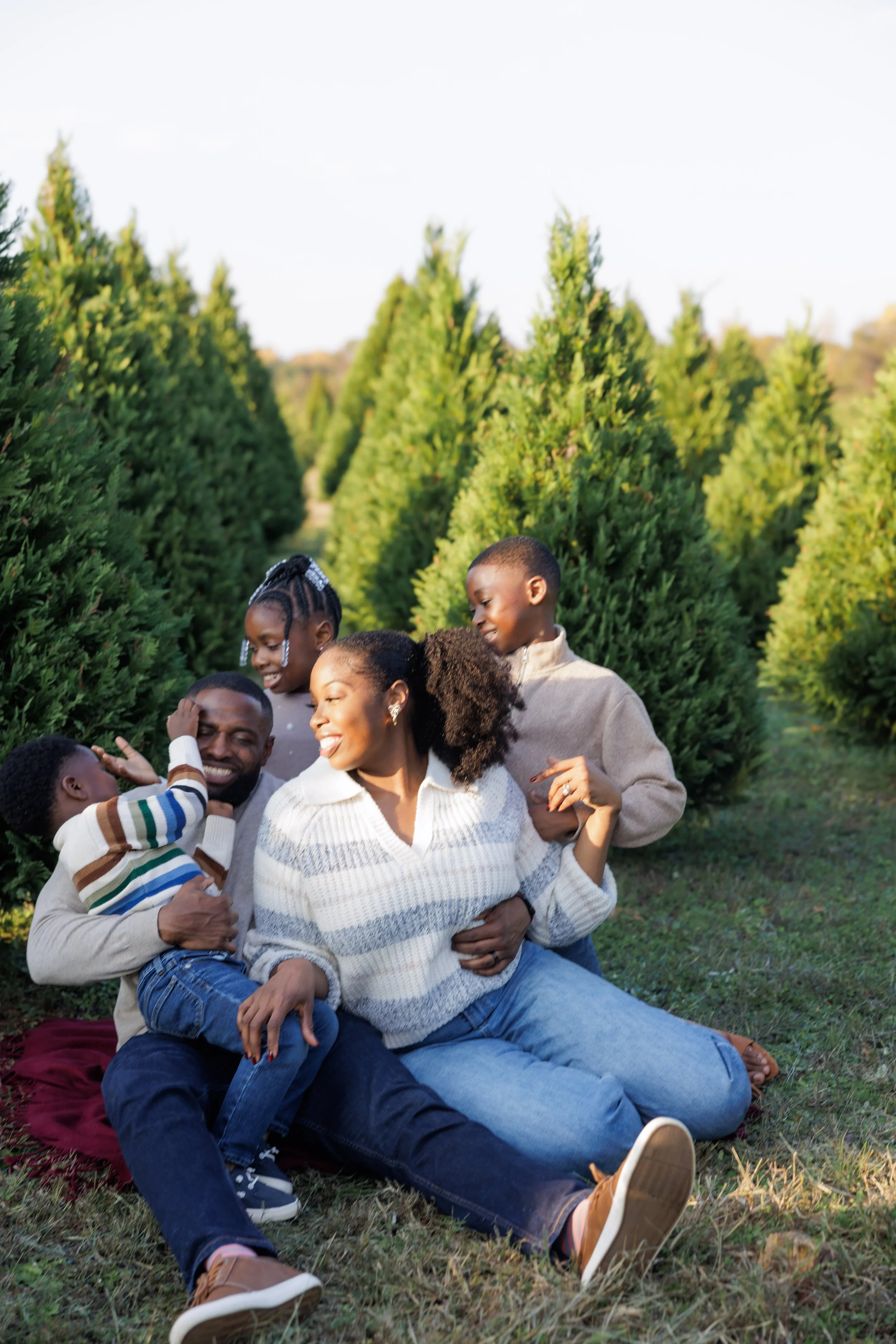 A happy family of five sitting and playing among evergreen trees outdoors during daytime.