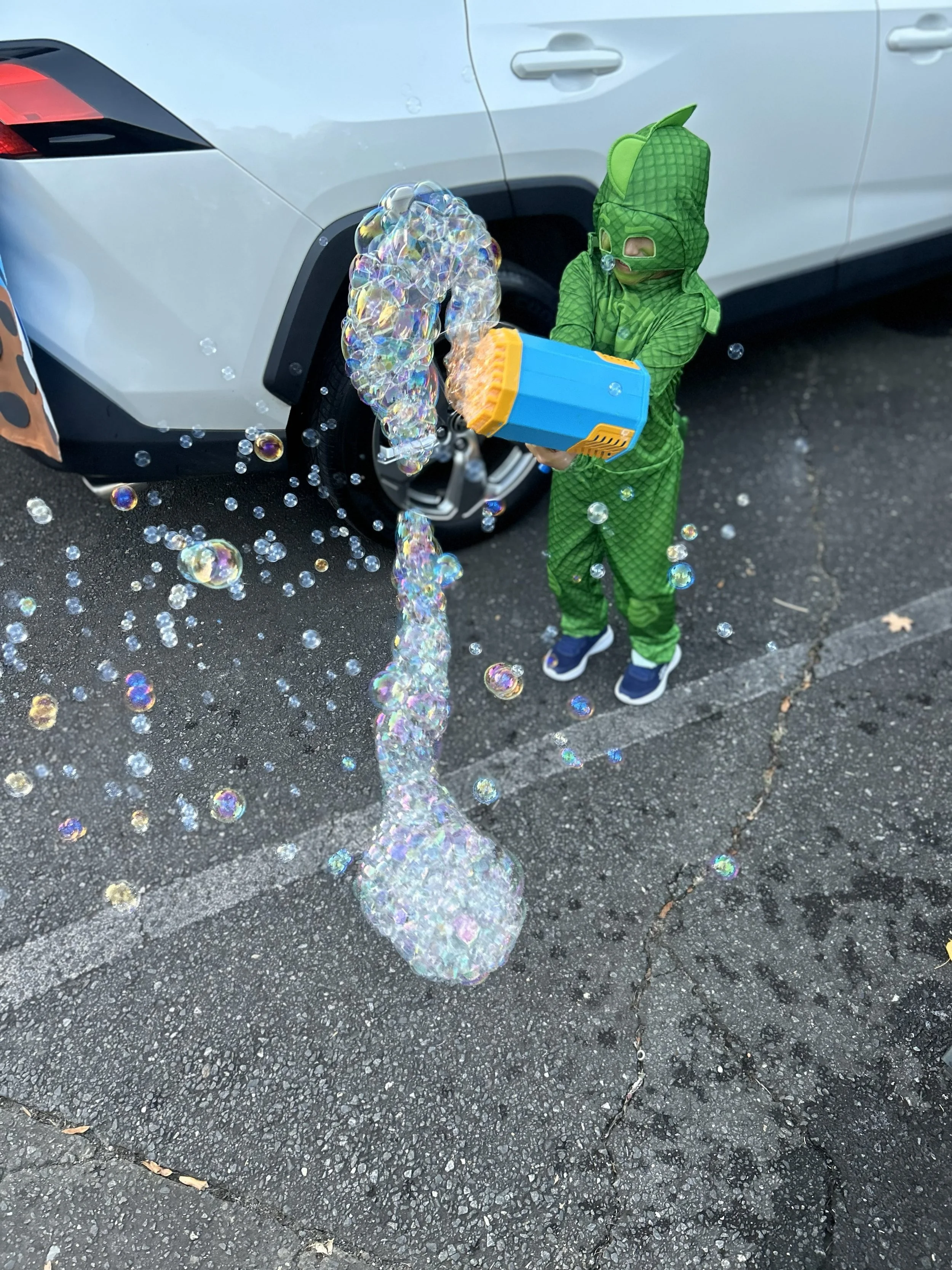 Child dressed in a green dinosaur costume blowing bubbles with a bubble gun in a parking lot next to a white vehicle.