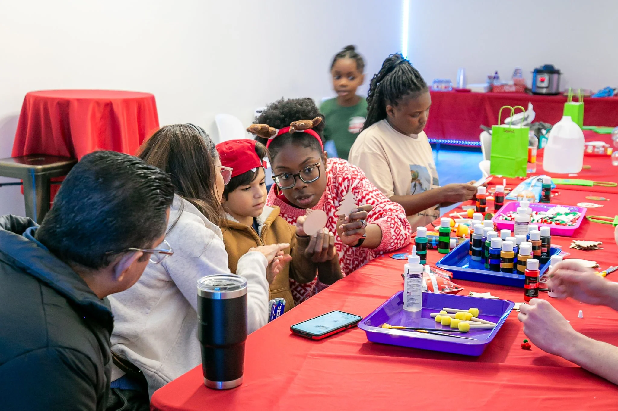 Children and adults participating in a craft activity at a holiday-themed event, with craft supplies on a red tablecloth.