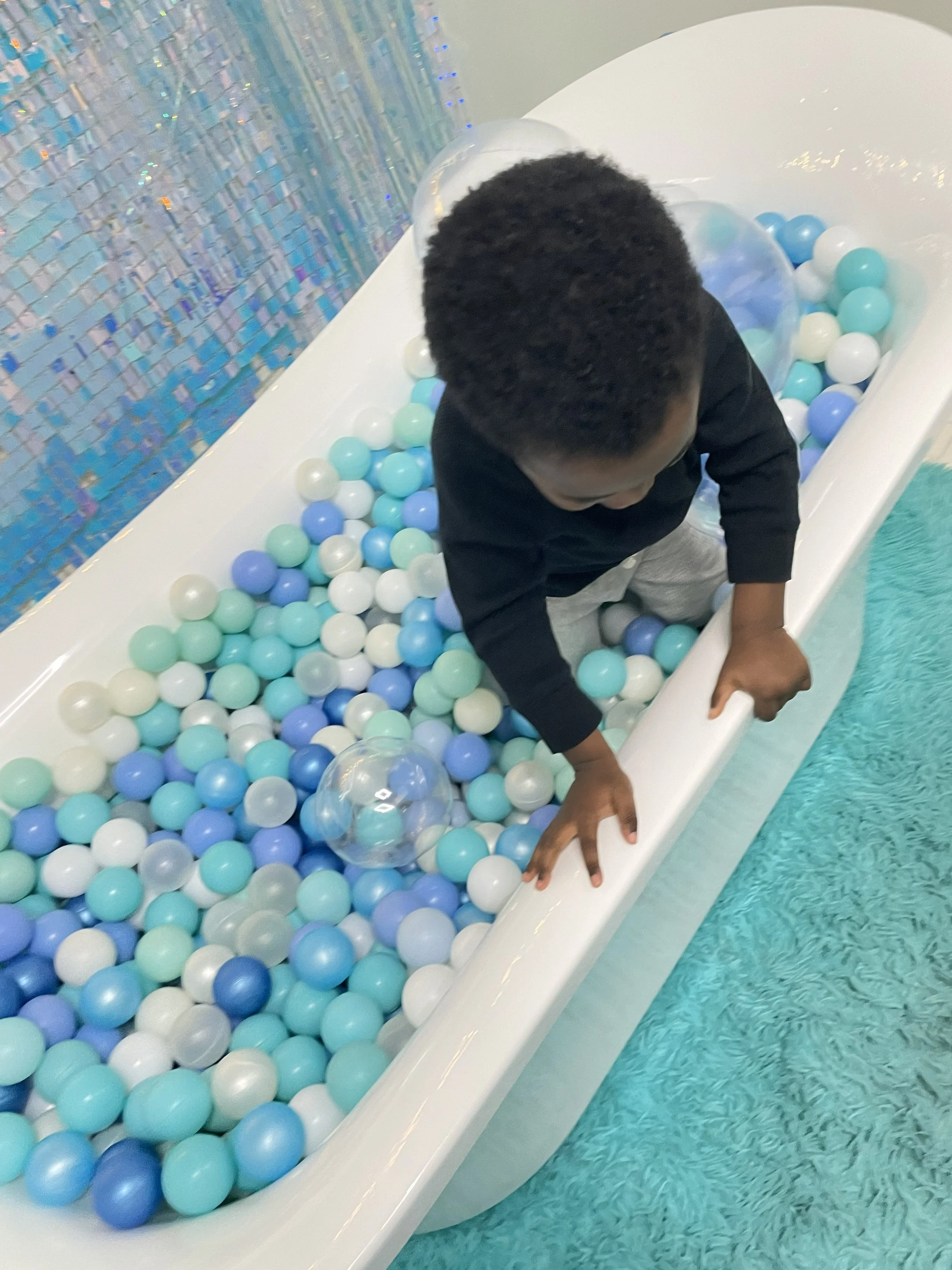 A young boy playing in a ball pit filled with blue, white, and mint green plastic balls, inside a room with a shiny, mosaic blue wall and a turquoise carpet.
