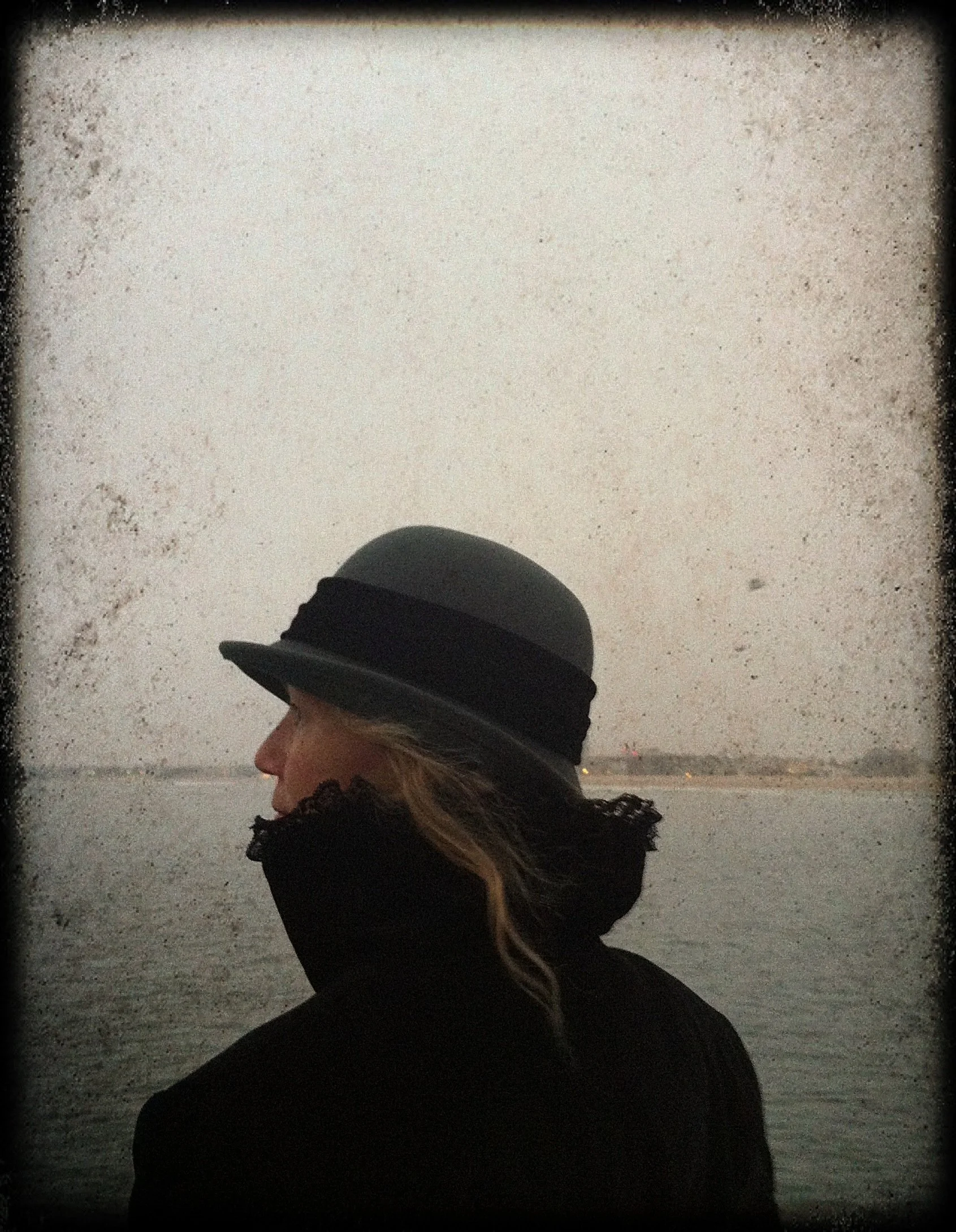 A woman with long hair, wearing a black coat and a black wide-brimmed hat, stands outside in moody weather overlooking a body of water with a distant shoreline.