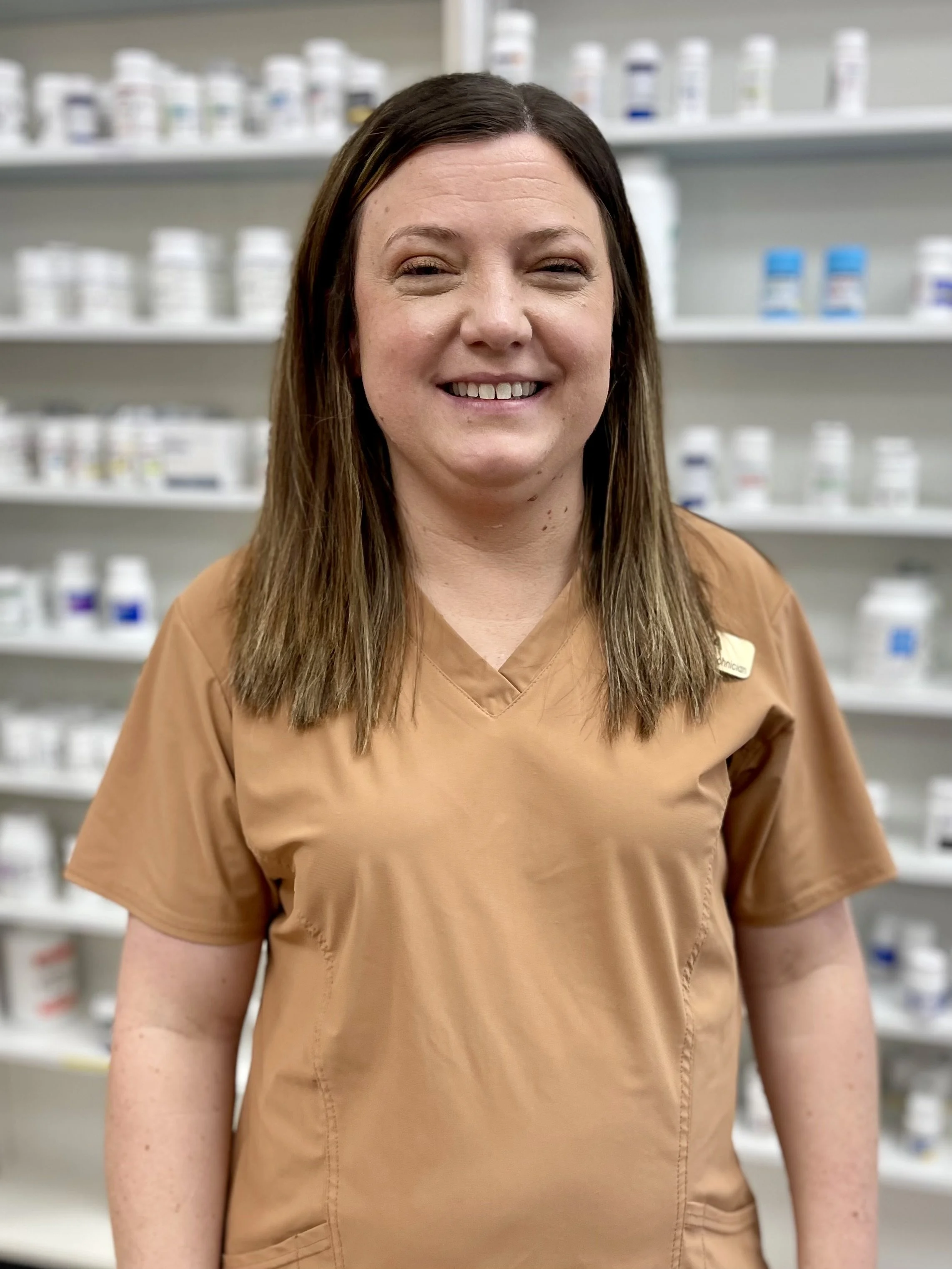 A smiling woman wearing tan medical scrubs standing in front of shelves with medication bottles.
