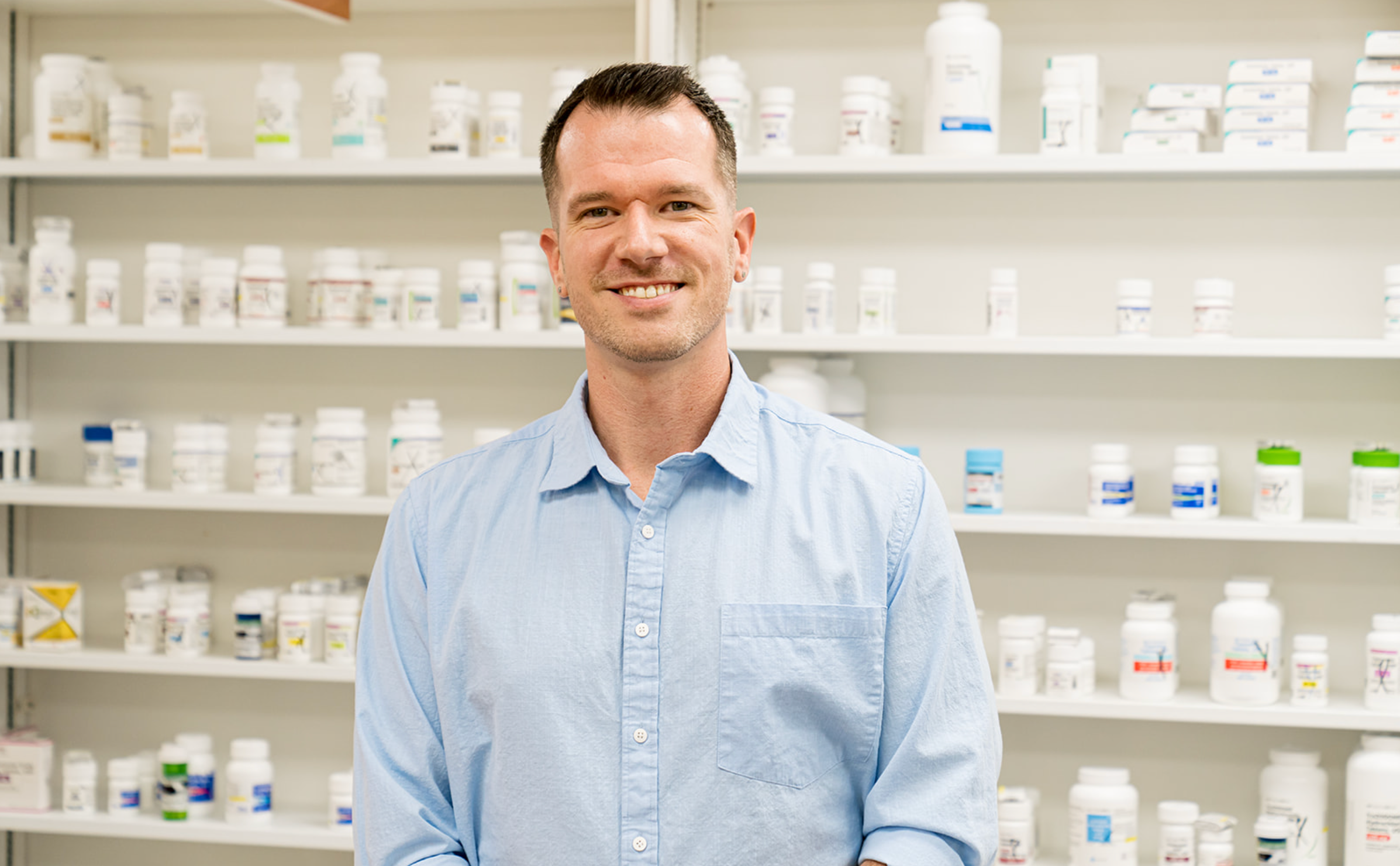 A man in a light blue shirt standing in front of shelves filled with medicine bottles in a pharmacy or medical supply store.