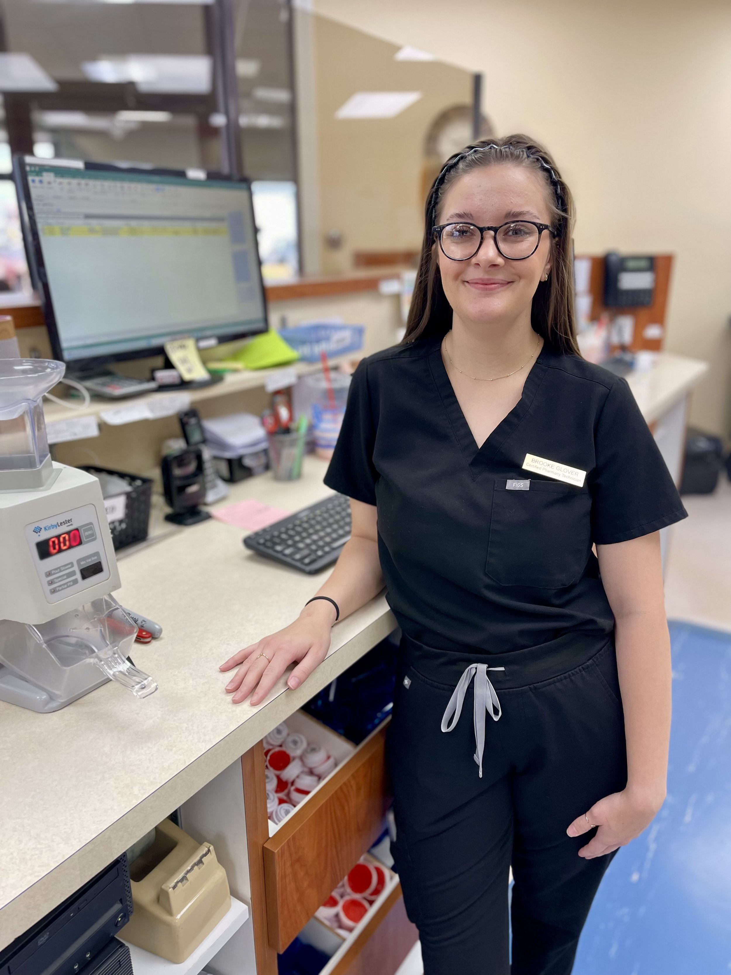 A smiling woman wearing glasses and a black uniform stands at a counter in a pharmacy, with medical supplies and a computer in the background.
