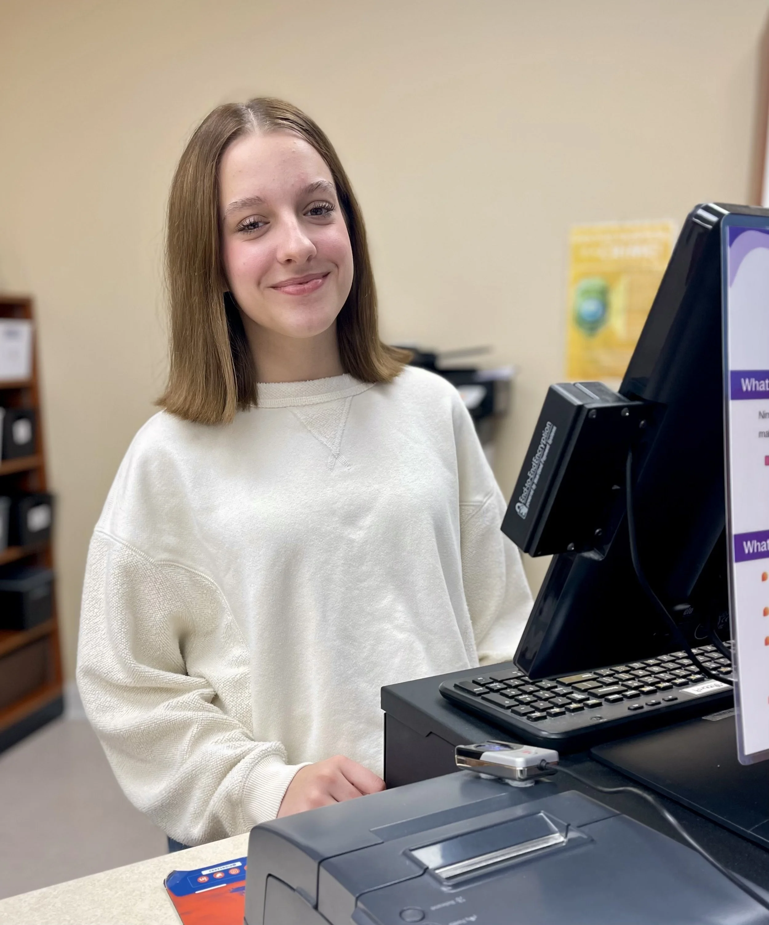A young woman with shoulder-length brown hair and a light-colored sweater smiling while standing at a checkout station in a store.