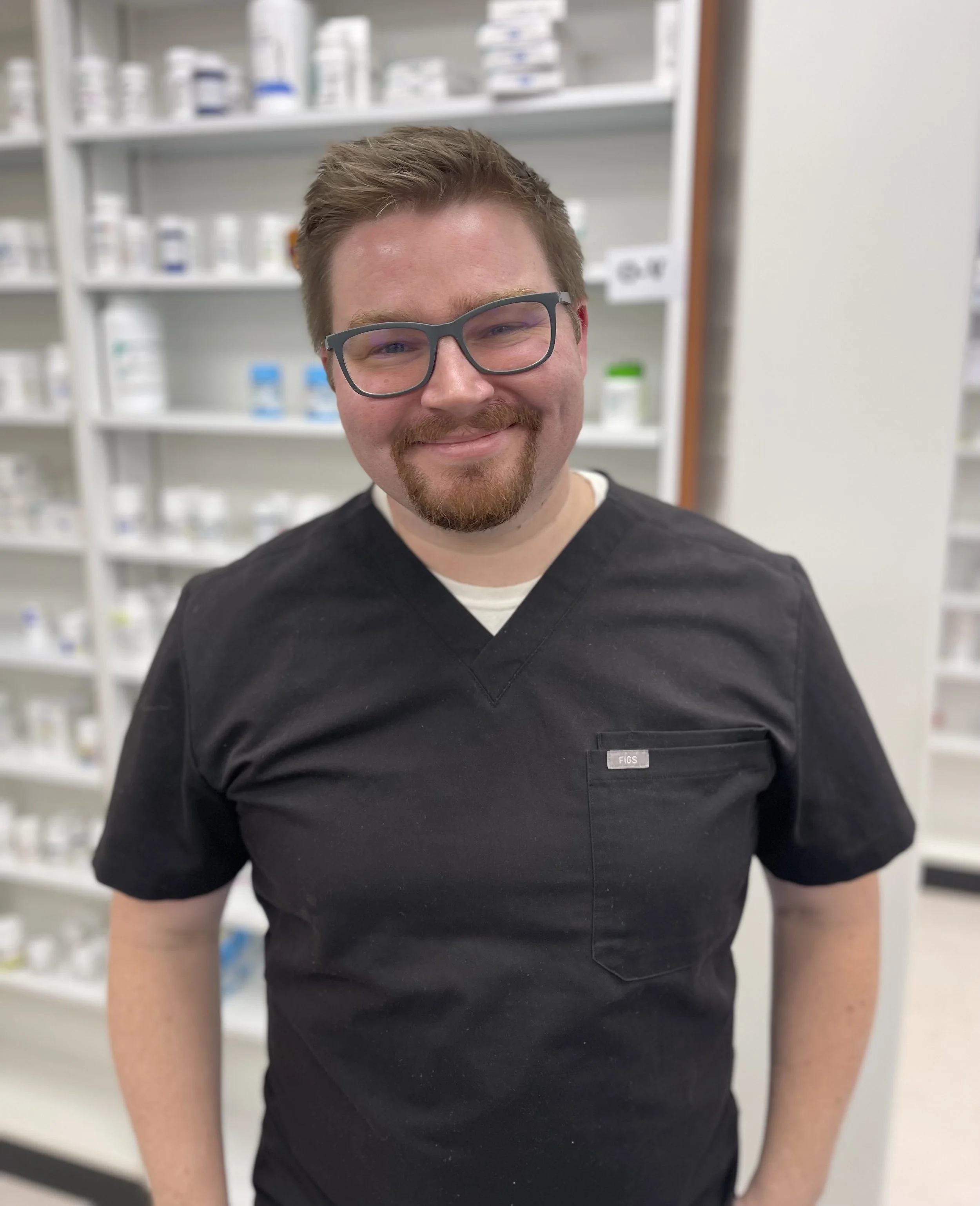 A smiling man with glasses and a beard wearing a black medical scrubs standing in front of shelves filled with medicine bottles and containers in a pharmacy.