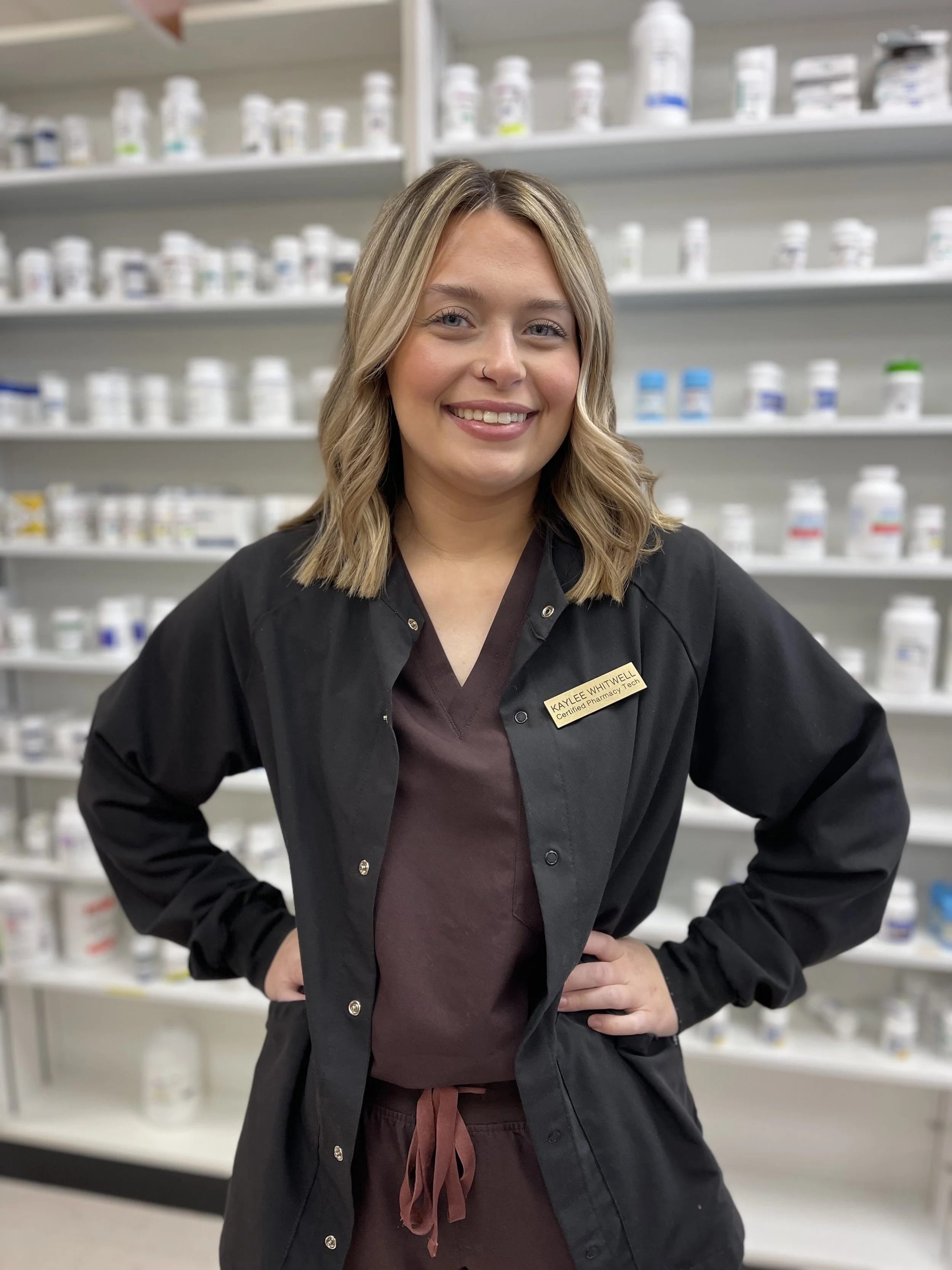 A smiling young woman with blonde hair and light blue eyes wearing black scrubs and a name tag in a pharmacy. Shelves of medication bottles are in the background.