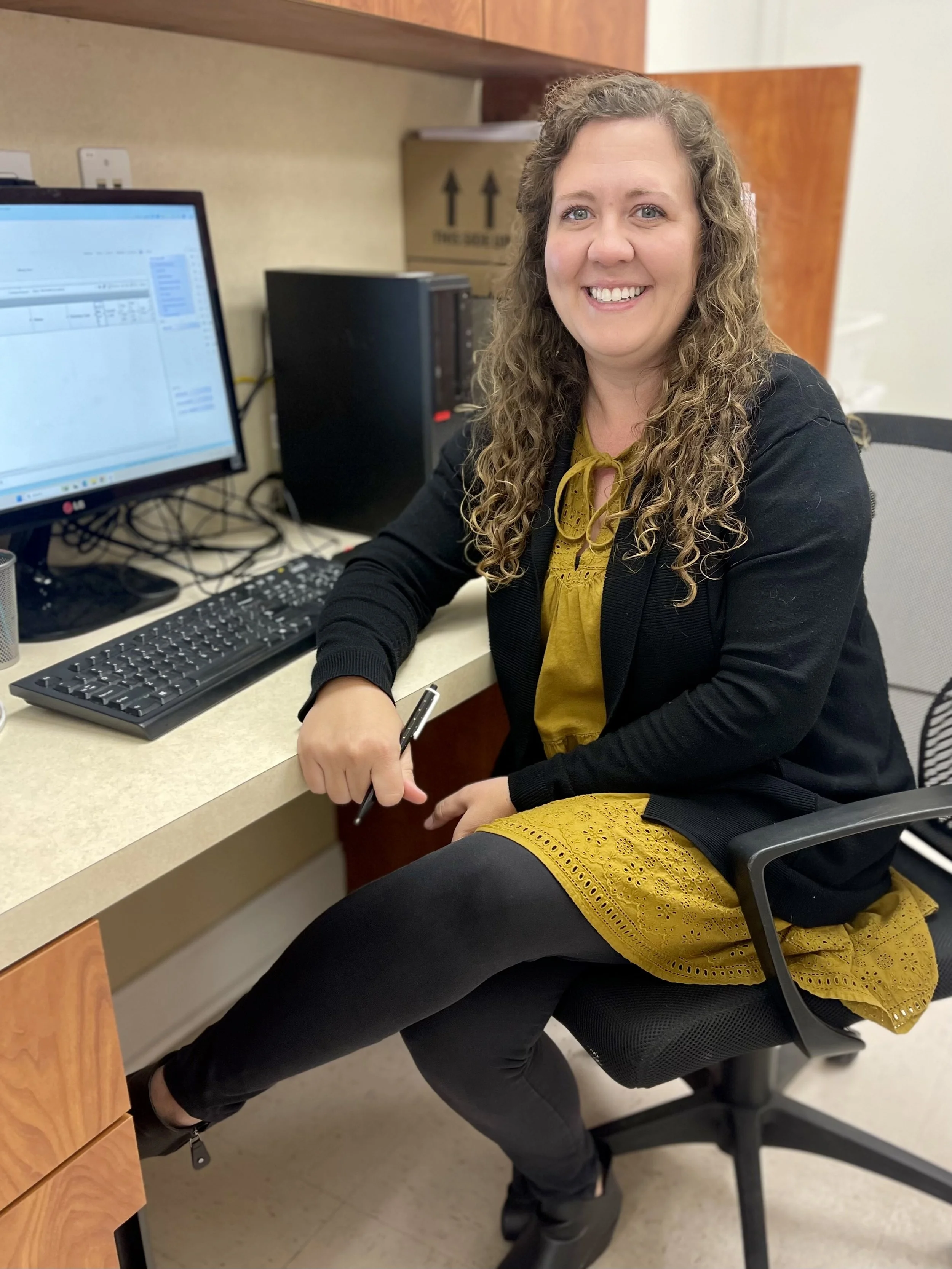 A woman sitting at an office desk smiling, with a computer, keyboard, and monitor visible. She has curly hair, is wearing a yellow dress and a black cardigan, and is holding a pen. There are cardboard boxes and office supplies in the background.