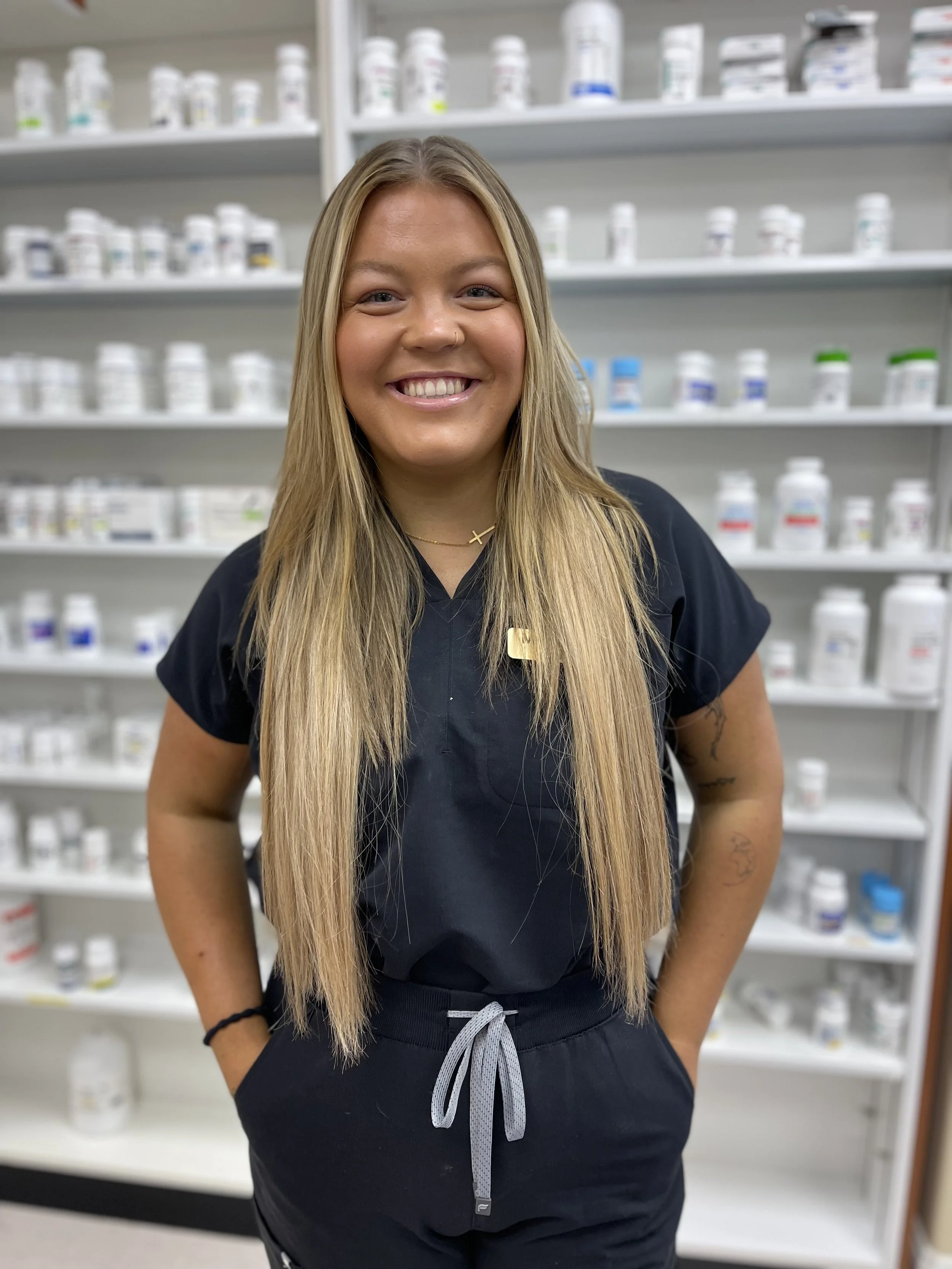 A young woman with long blonde hair smiling in front of pharmacy shelves filled with bottles.