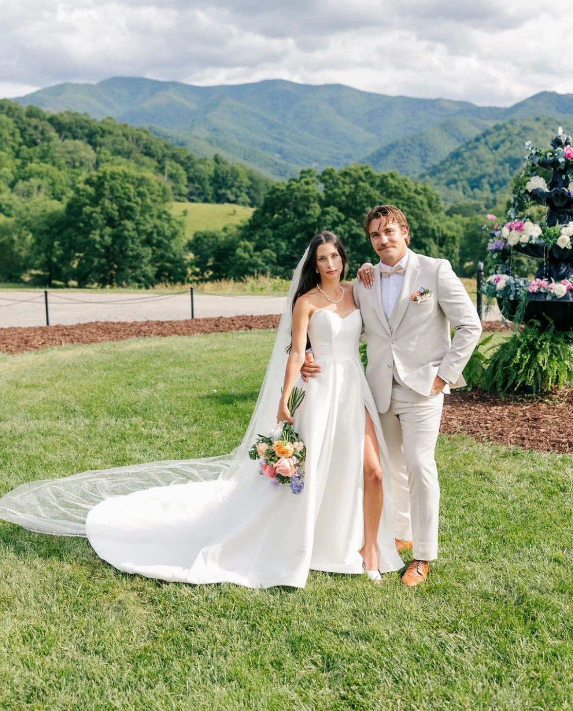 A bride and groom standing together outdoors during their wedding, with mountains and green trees in the background. The bride is wearing a white wedding dress with a thigh-high slit, holding a bouquet of flowers. The groom is in a light-colored suit