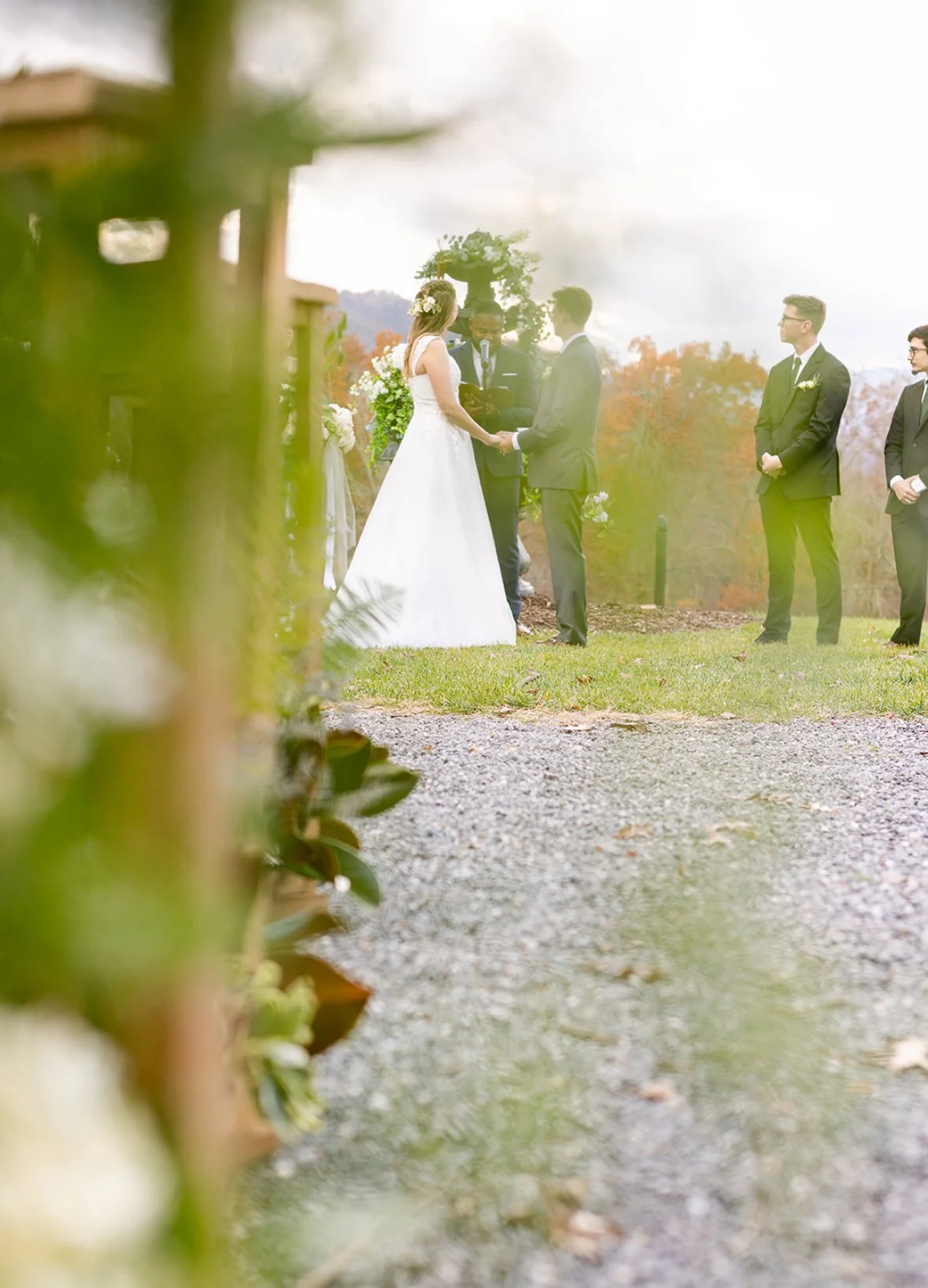 A couple getting married outdoors, standing before an officiant, with bridesmaids and groomsmen standing beside them. The scene is framed by blurred greenery in the foreground, with a natural background of trees and a cloudy sky.