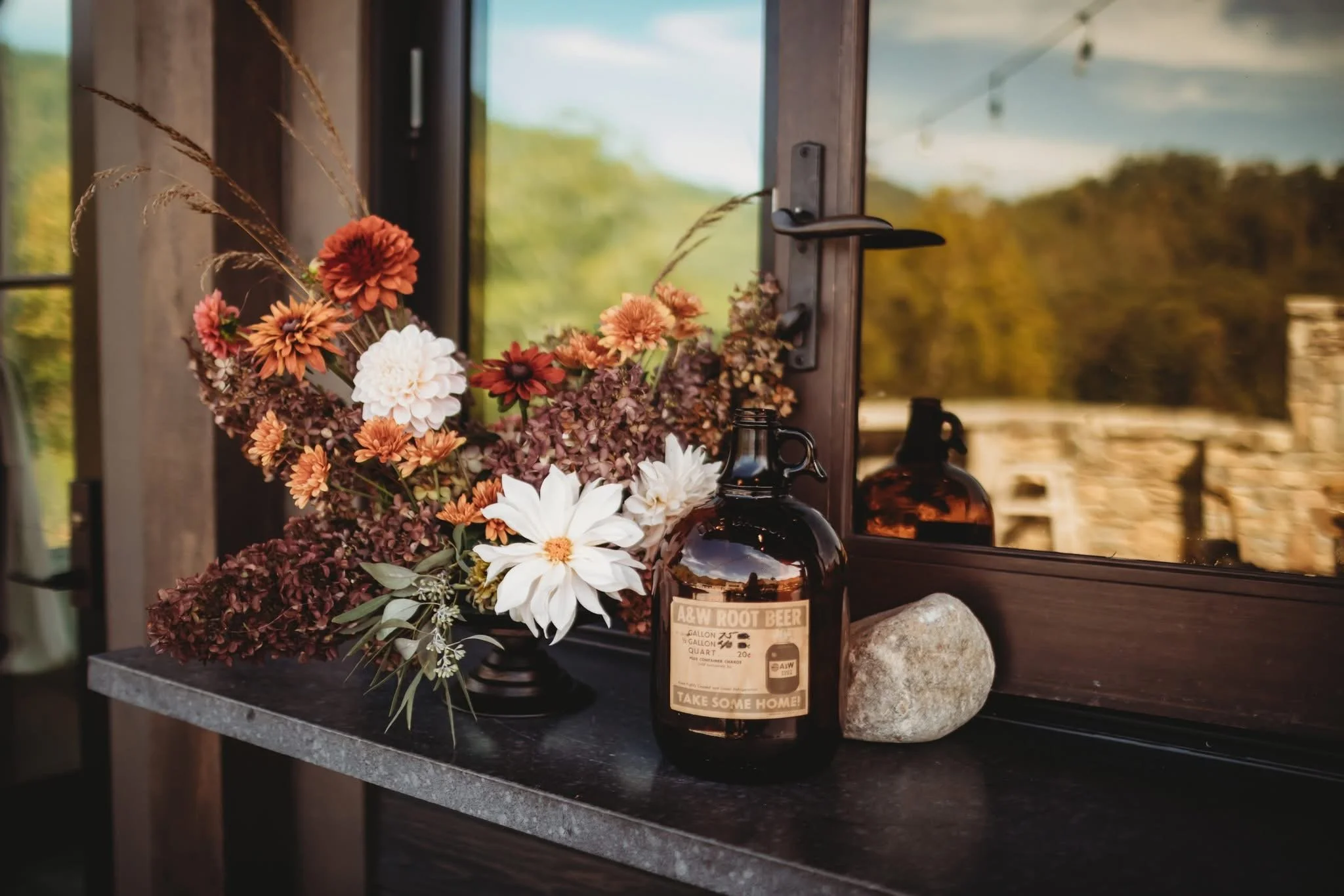 A bouquet of mixed flowers with shades of white, pink, red, and purple, placed on a dark table. Next to it, a dark glass bottle with a label reading 'A&W Root Beer' and a large rock. The background shows a window with a reflection of the outdoors and
