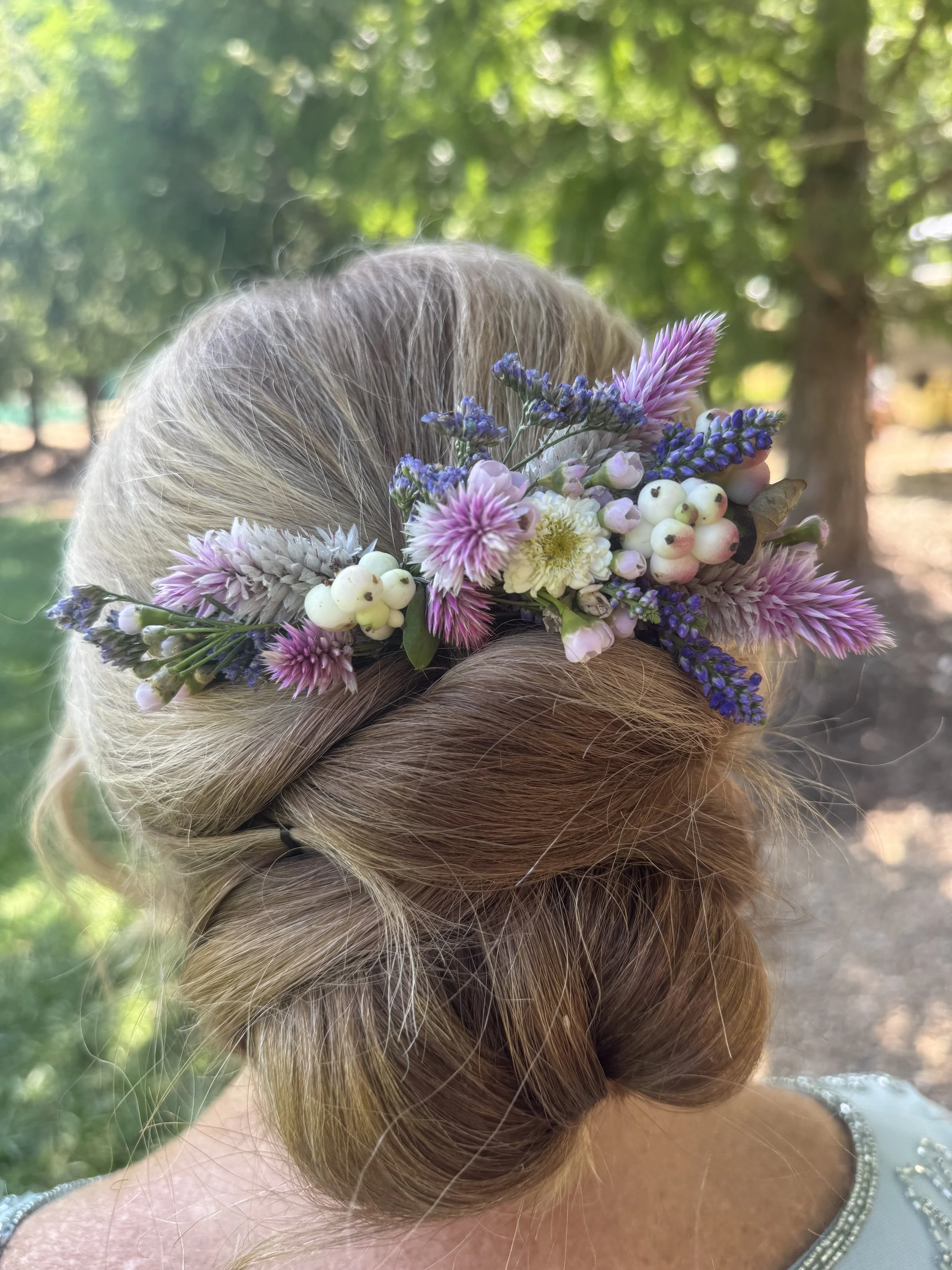 Close-up of a woman with blonde hair styled in an elegant updo, wearing a floral crown made of pink, purple, white flowers, and white berries, outdoors with trees and sunlight in the background.
