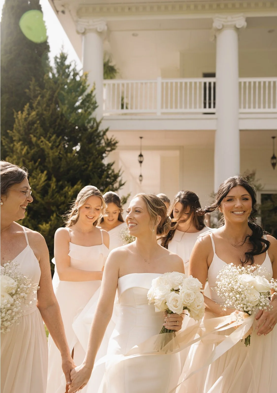 A group of women smiling and holding bouquets of white flowers, dressed in white gowns, standing outdoors in front of a large white house with pillars, with sunlight and greenery in the background.