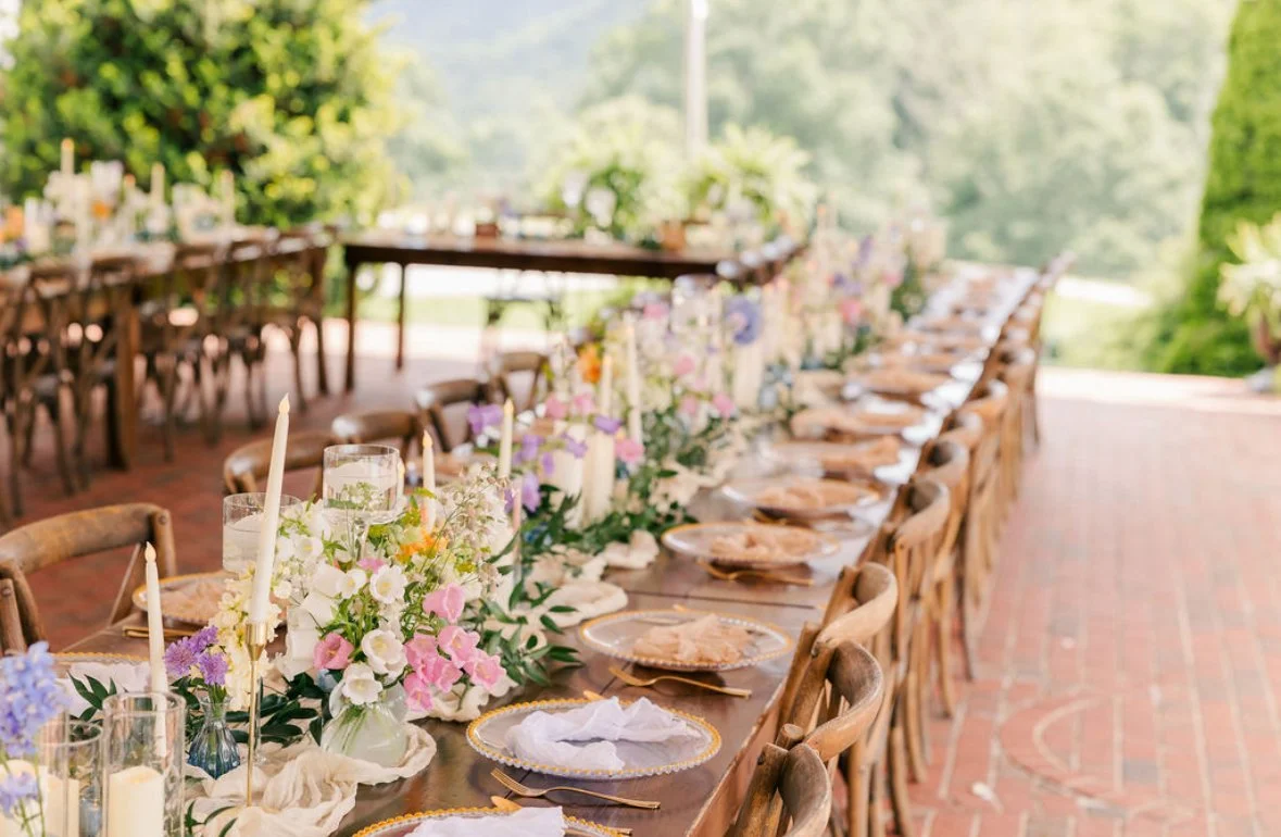 Long outdoor dining table decorated for a celebration, with floral centerpieces, candles, and place settings, set in a lush green garden.