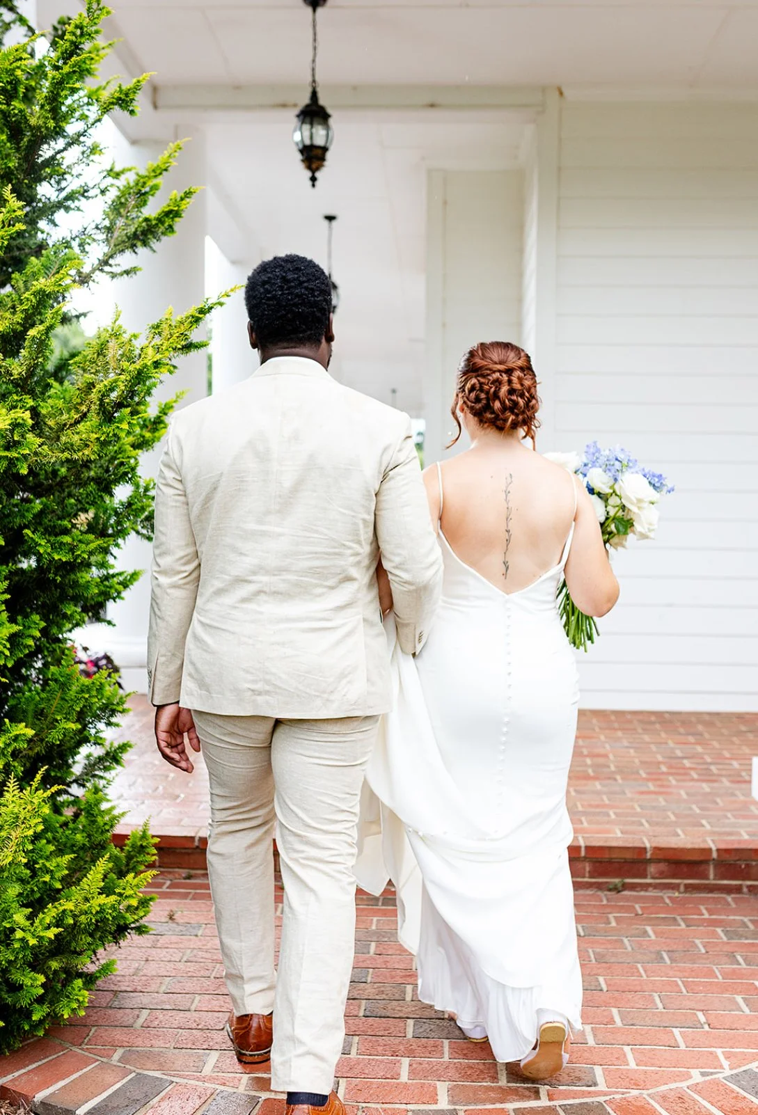 A couple dressed in wedding attire walking away on a brick pathway in front of a white house, with a woman holding a bouquet of flowers.
