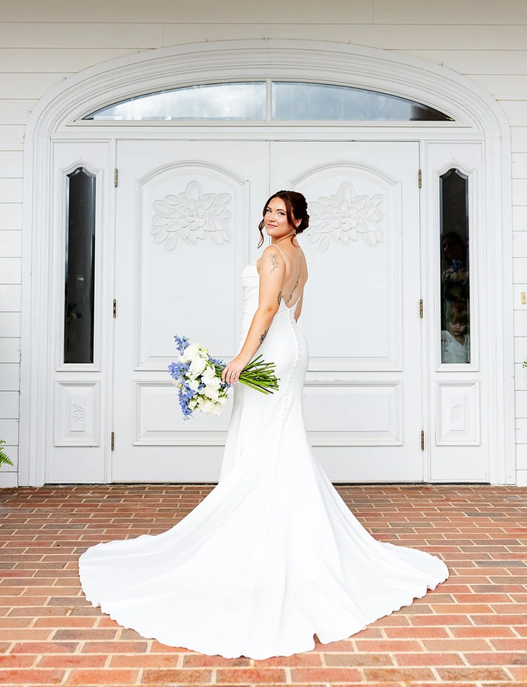 A bride in a white wedding gown holding a bouquet of white and blue flowers, standing in front of a white door with decorative carvings, brick paver walkway.