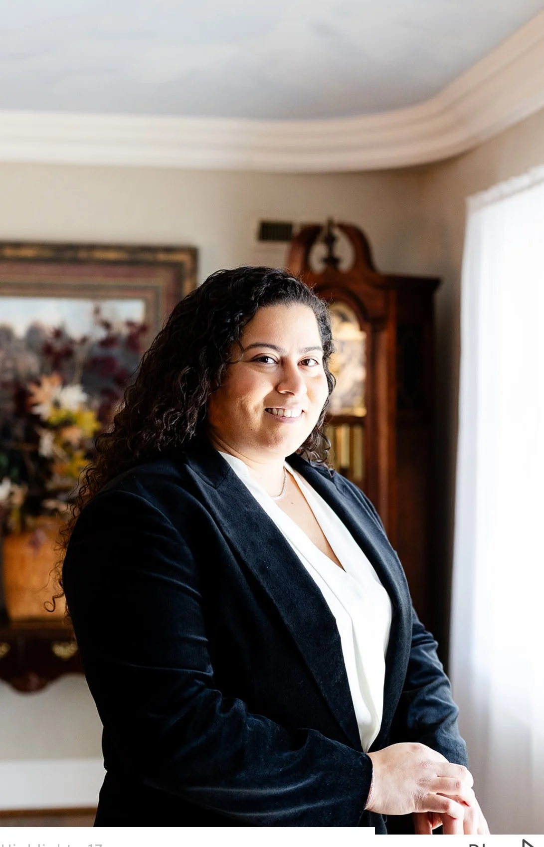A woman with curly dark hair smiling, wearing a black blazer and white blouse, standing indoors in front of a wooden cabinet and floral painting, with natural light coming from a window.