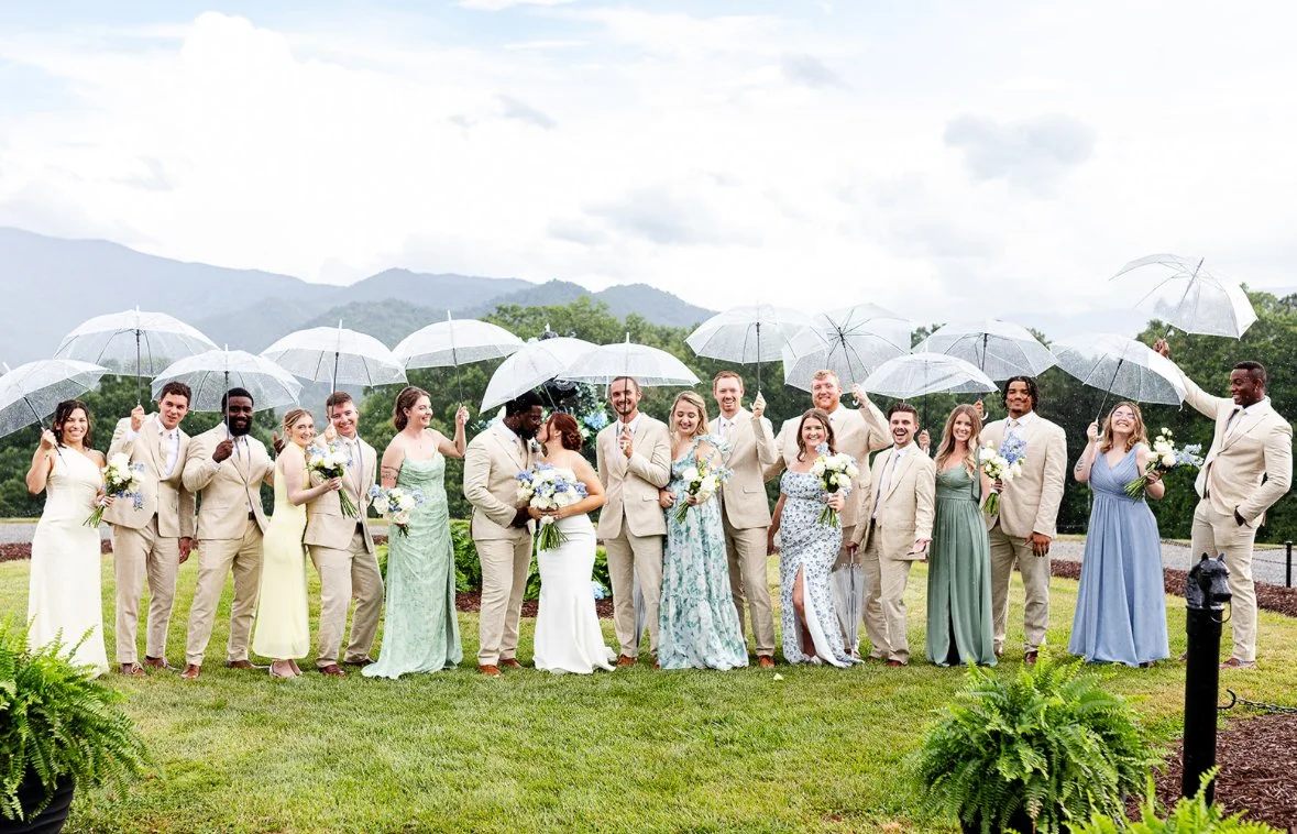A wedding party standing outdoors on a grassy area, holding transparent umbrellas in a light rain, with a scenic mountain background. The group includes bridesmaids and groomsmen dressed in pastel and neutral colors, smiling and celebrating.