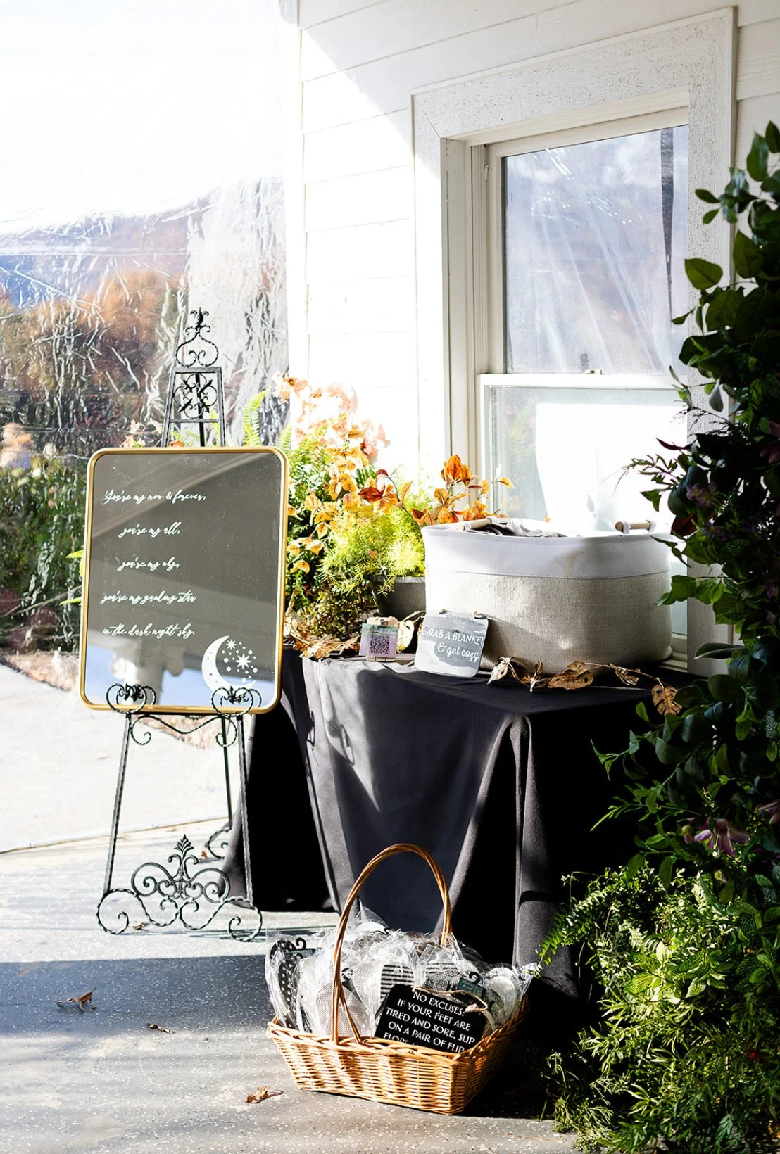 Indoor scene with a mirror on a stand, a table covered with a black cloth, a basket filled with ice and bottles, and a variety of plants and flowers near a window.