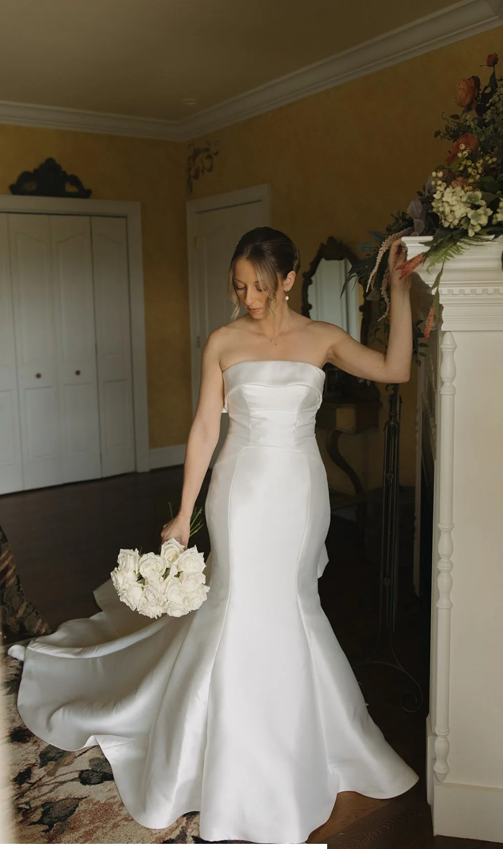 A bride in a strapless white wedding gown holding a bouquet of white roses, standing indoors near a fireplace.