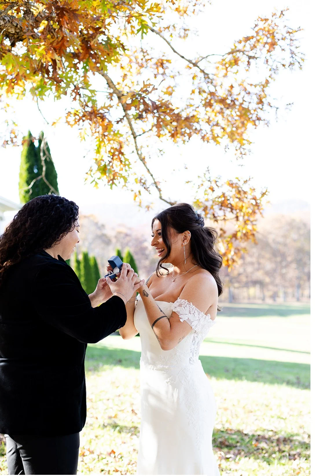 A woman in a white wedding dress receiving a ring from a person in black clothing outdoors, with fall-colored leaves and trees in the background, smiling and happy.