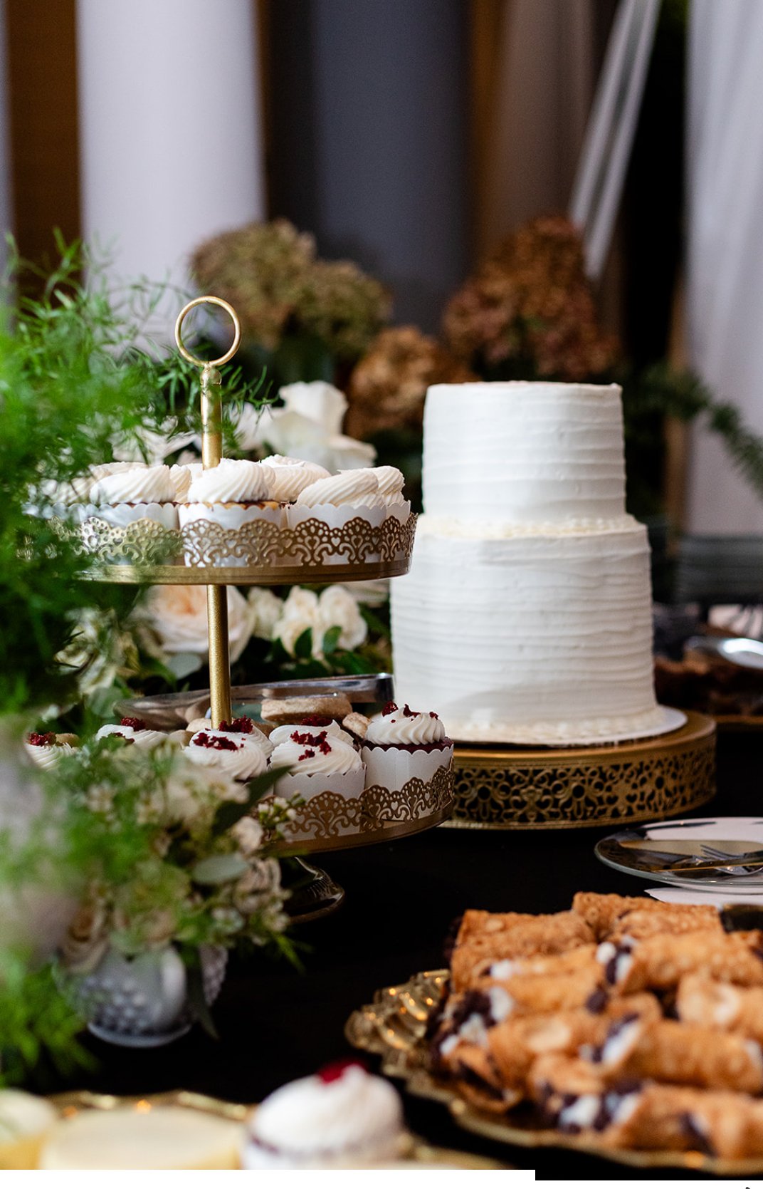A dessert table with a two-tiered white frosted cake, a tiered tray of cupcakes with white frosting, and assorted cookies, all surrounded by greenery and floral arrangements in a decorated setting.
