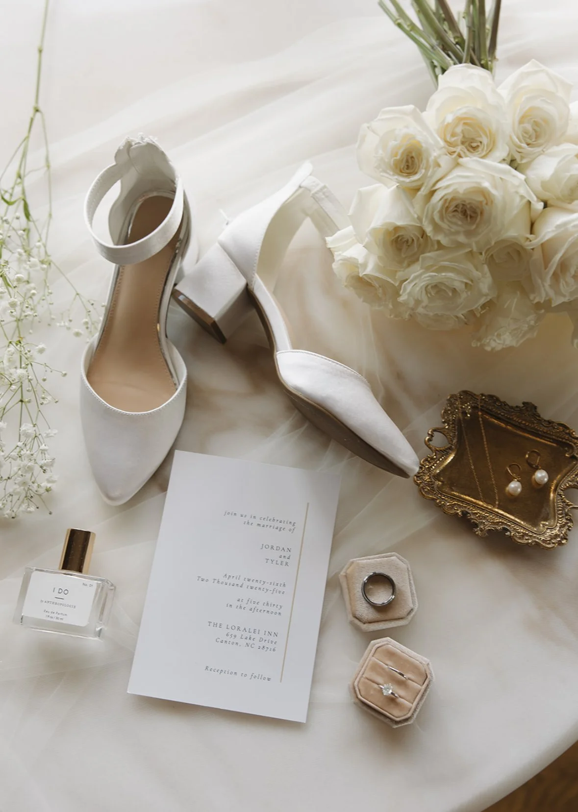 A bouquet of white roses, a pair of white high-heeled shoes, a wedding invitation, pearl earrings, a wedding ring in a velvet box, a perfume bottle, and a gold jewelry tray are arranged on a white surface.