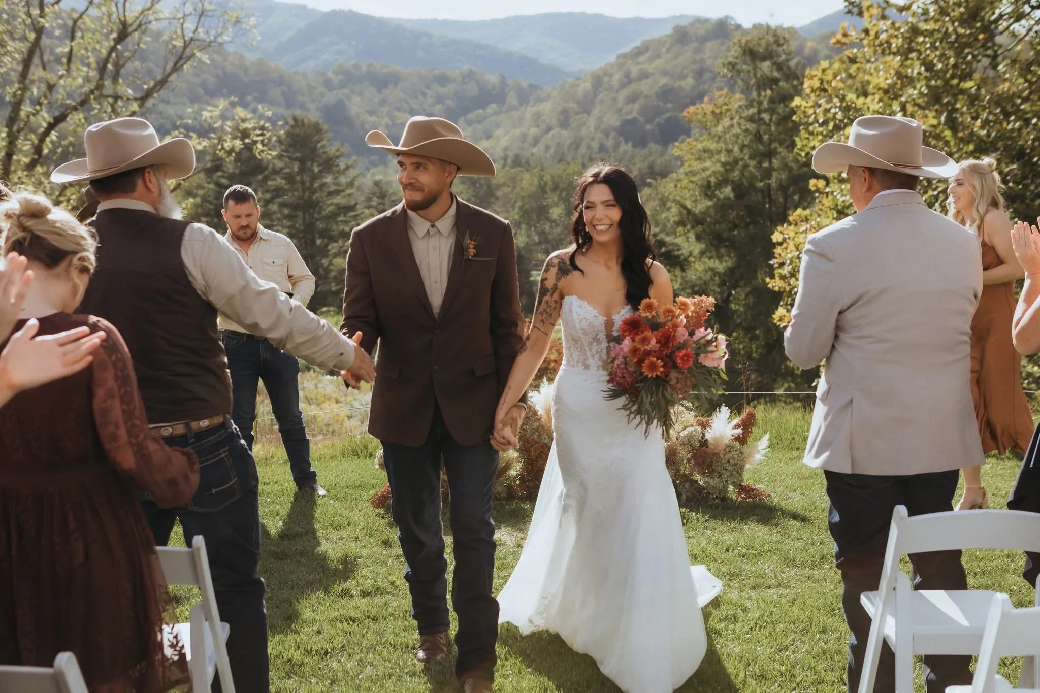 Bride and groom holding hands and walking outdoors at their wedding ceremony, surrounded by guests, with scenic mountains and trees in the background.