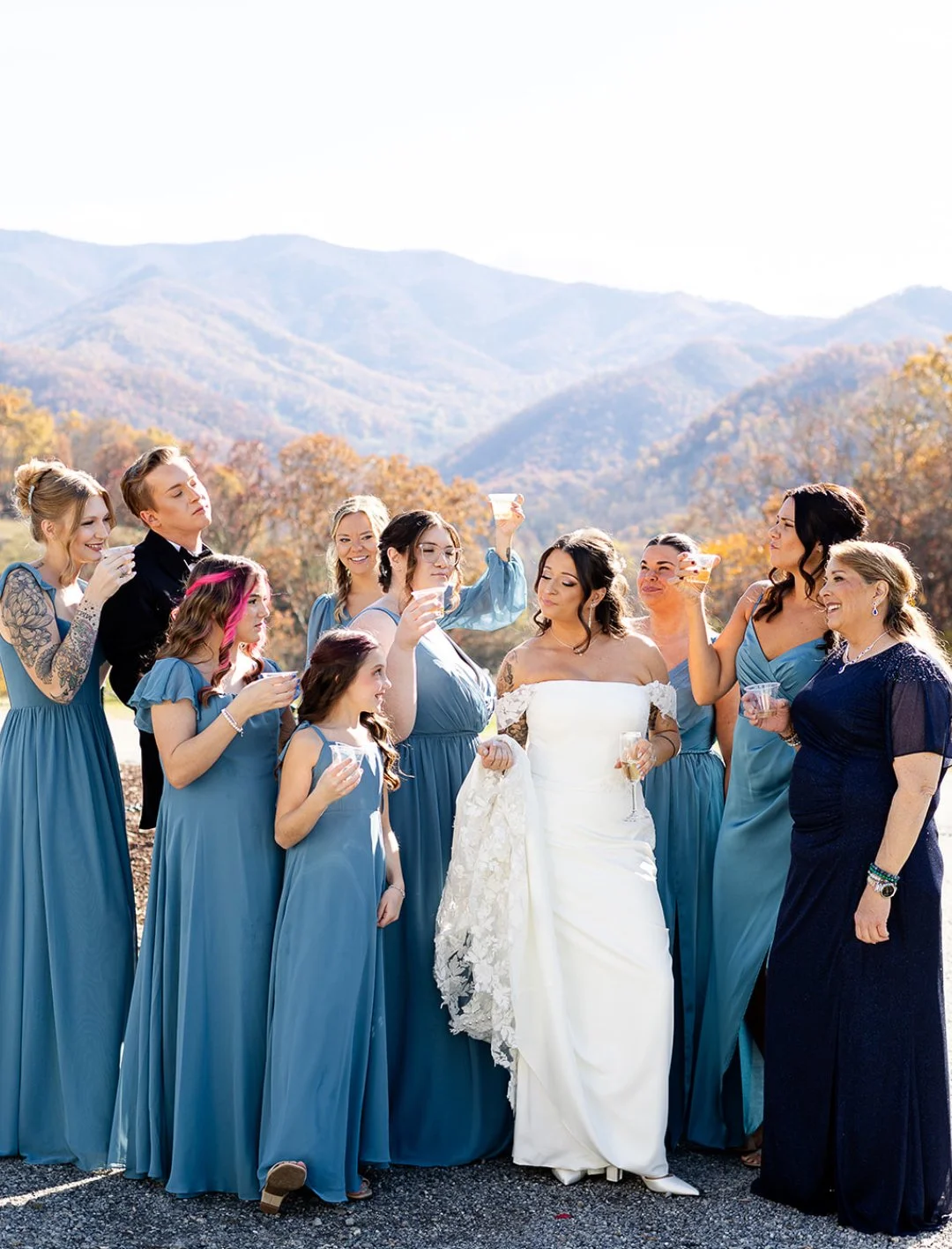 A group of women in matching blue dresses and a bride in a wedding gown holding glasses of champagne, enjoying an outdoor celebration with mountains and autumn trees in the background.