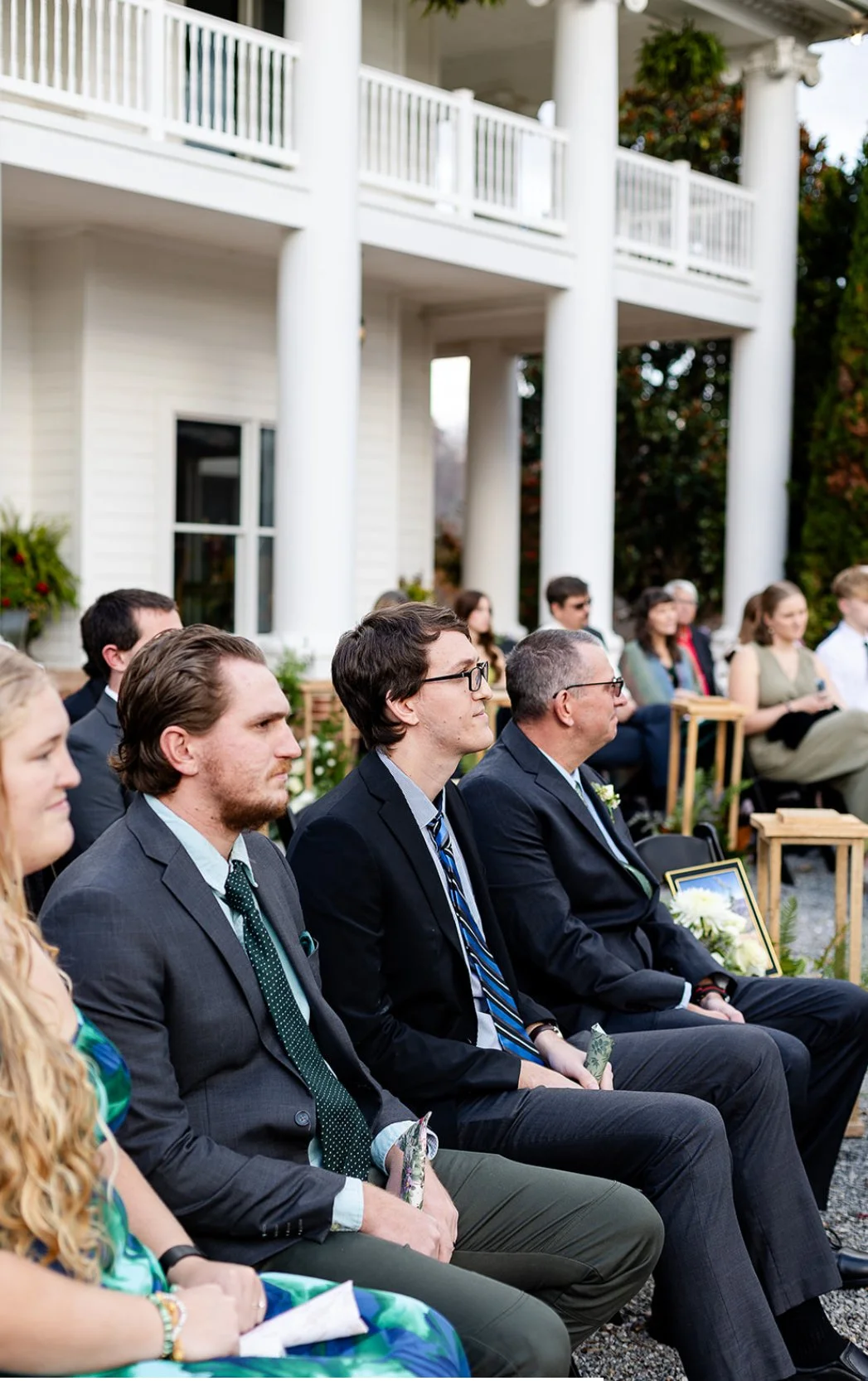 People attending outdoor wedding ceremony seated in front of a white house with large columns and a balcony, dressed in formal and semi-formal attire.
