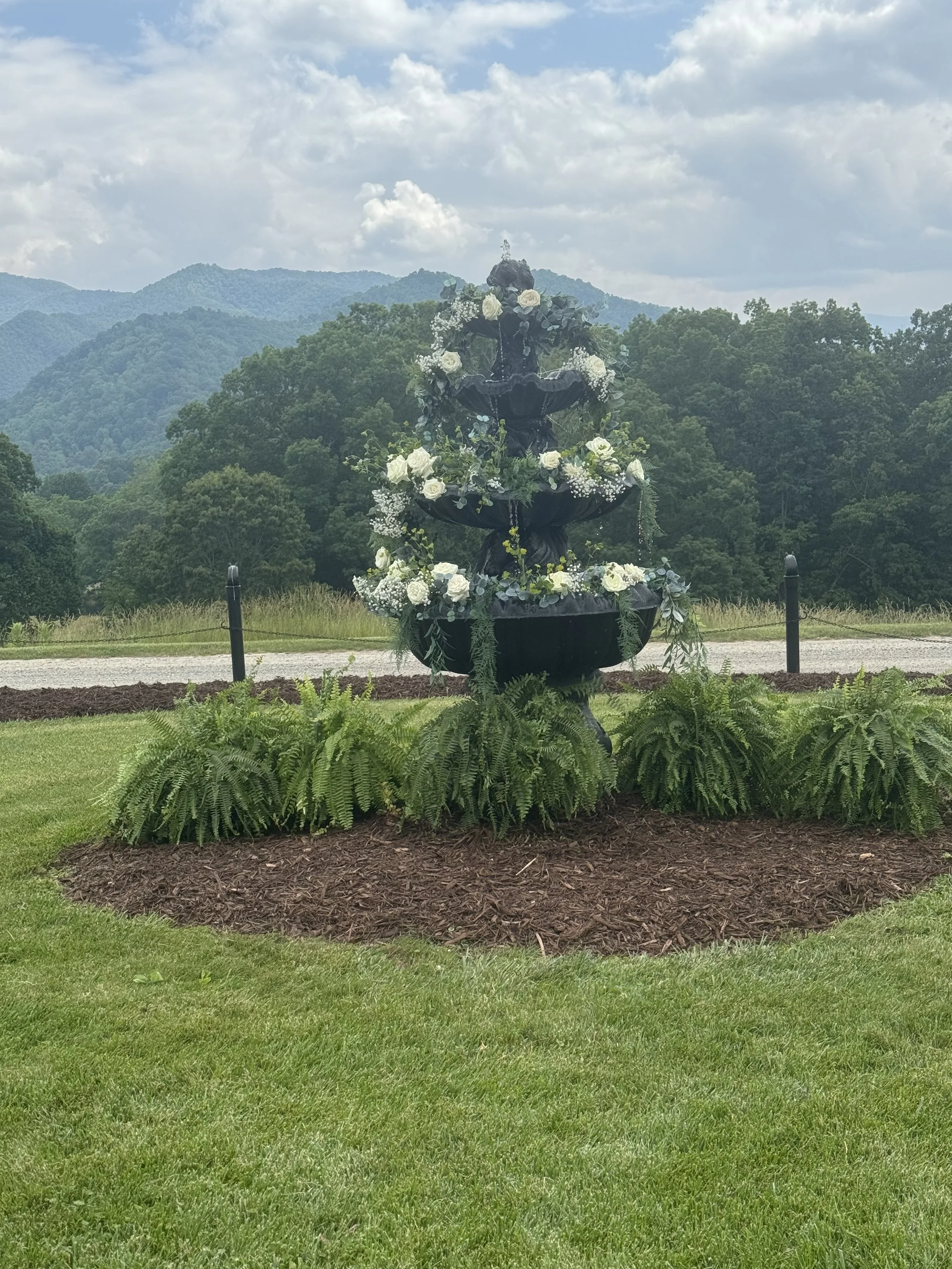 A decorative outdoor fountain covered with white flowers and greenery, surrounded by a landscaped garden with lush green grass and ferns, with a backdrop of mountains and a cloudy sky.