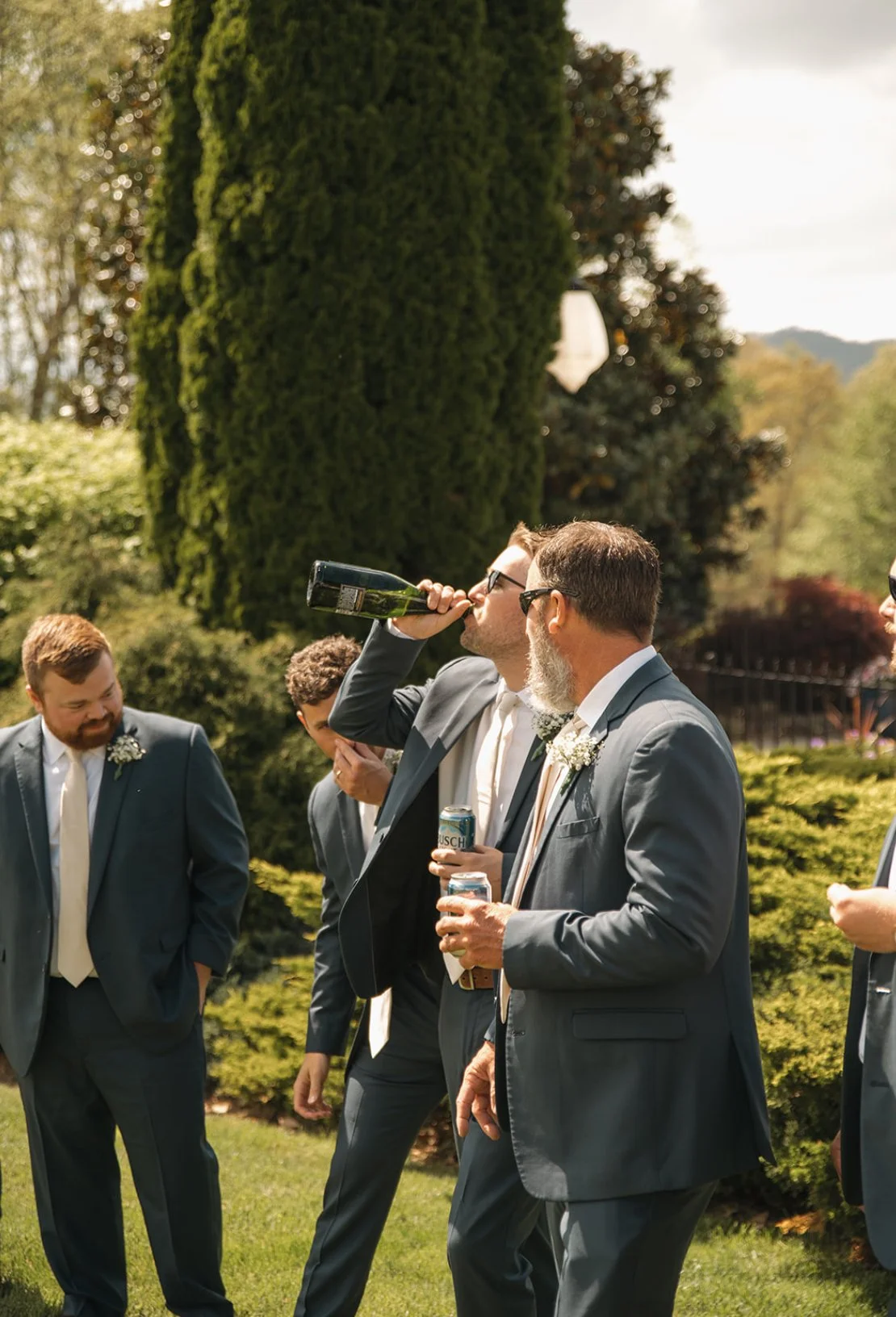 Group of men in suits at an outdoor wedding, with one man drinking from a bottle and others holding drinks.