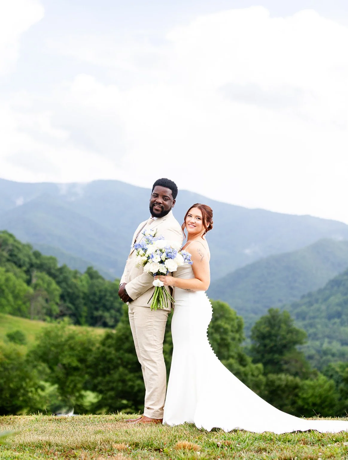 A couple dressed in wedding attire standing outdoors with mountains and green trees in the background, holding a bouquet of flowers.