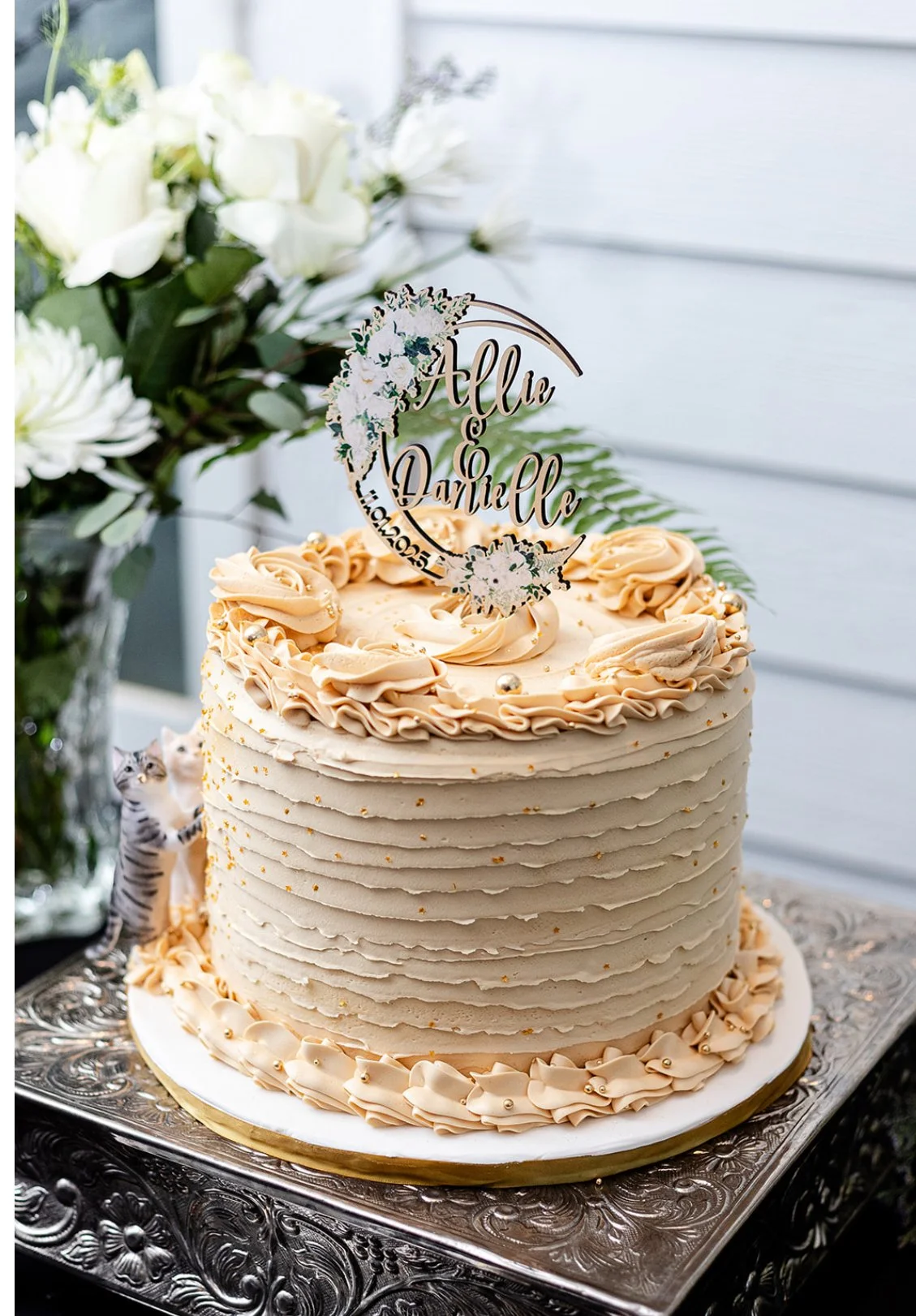 A decorated wedding cake with white and peach frosting, topped with a custom cake topper that reads 'Allie & Danielle' and the date '10.20.23'. The cake has rosette and piped designs, gold accents, and is displayed on an ornate silver tray with a pur