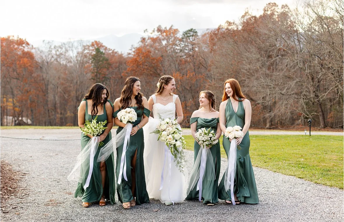 A bride and four bridesmaids walking outdoors on a gravel path with autumn trees in the background, all smiling and holding bouquets.